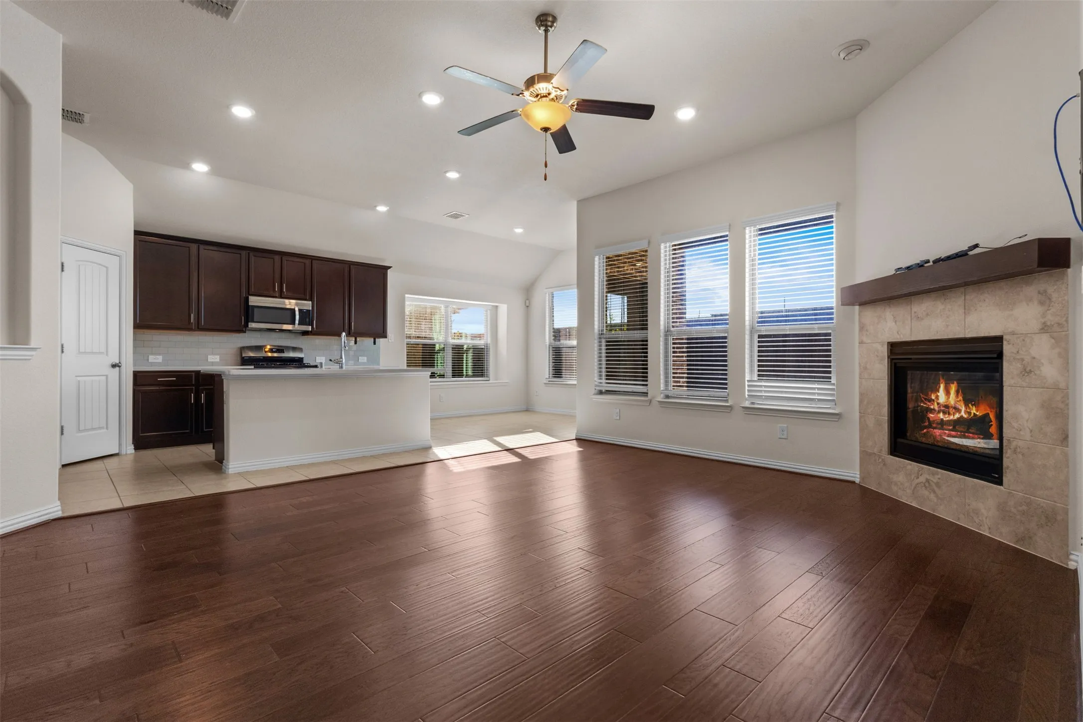 Vaulted ceiling with recessed lighting and ceiling fan, wood-finish flooring, tiled fireplace with wood mantel, kitchen featuring dark wood cabinetry and stainless steel appliances, and multiple windows with blinds