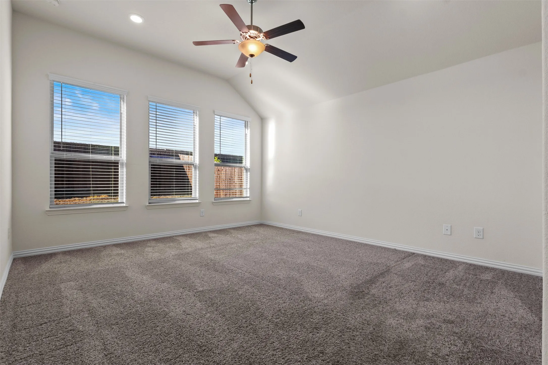 Vaulted ceiling, three windows with blinds, ceiling fan with integrated lighting, recessed lighting, and neutral carpet