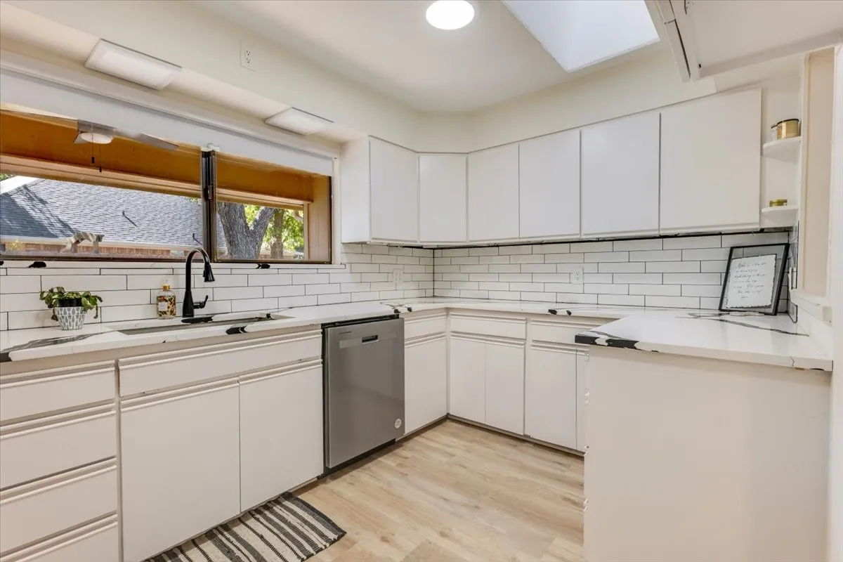 White cabinetry with black hardware, subway tile backsplash, stainless steel dishwasher, wood-finish flooring, and a skylight