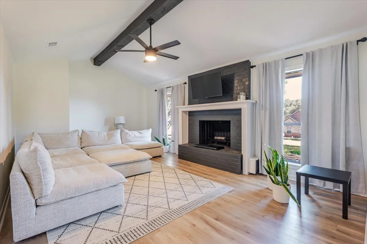 Vaulted ceiling with exposed beam, ceiling fan, wood-finish flooring, brick fireplace with mantel, and window with natural light