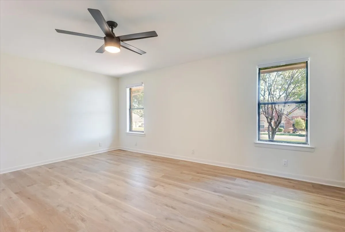 Light wood-finish flooring, two windows with white trim, and a ceiling fan with integrated lighting