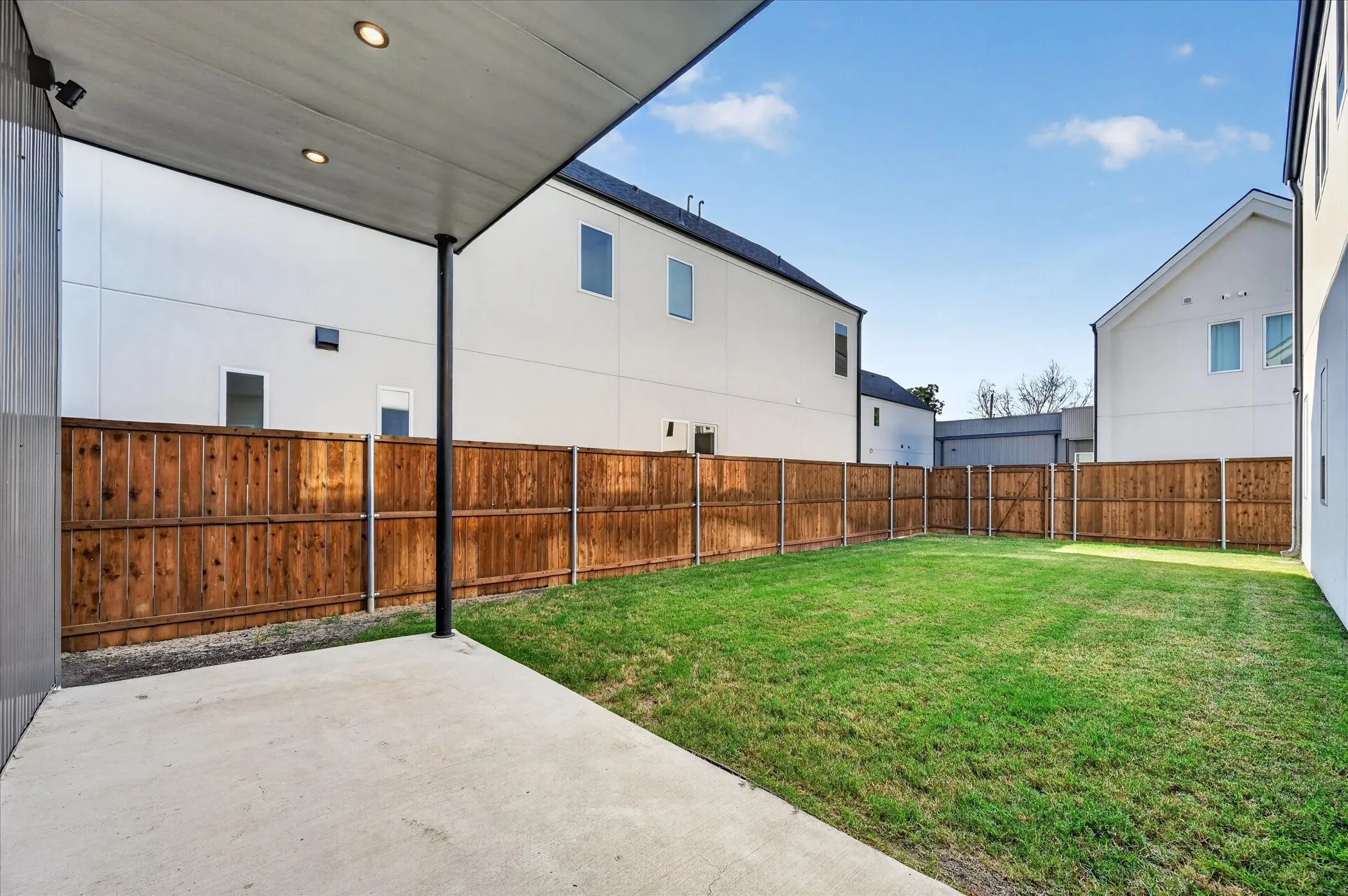 Expansive backyard featuring a manicured lawn, full perimeter wood fencing, and a concrete patio with an overhead cover and recessed lighting