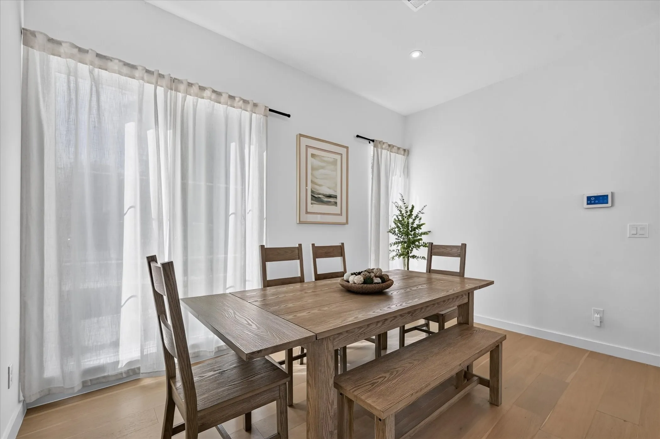 Bright dining area showcasing wood-finish flooring, a substantial wood dining table with seating for six, and large windows with sheer curtains providing natural light