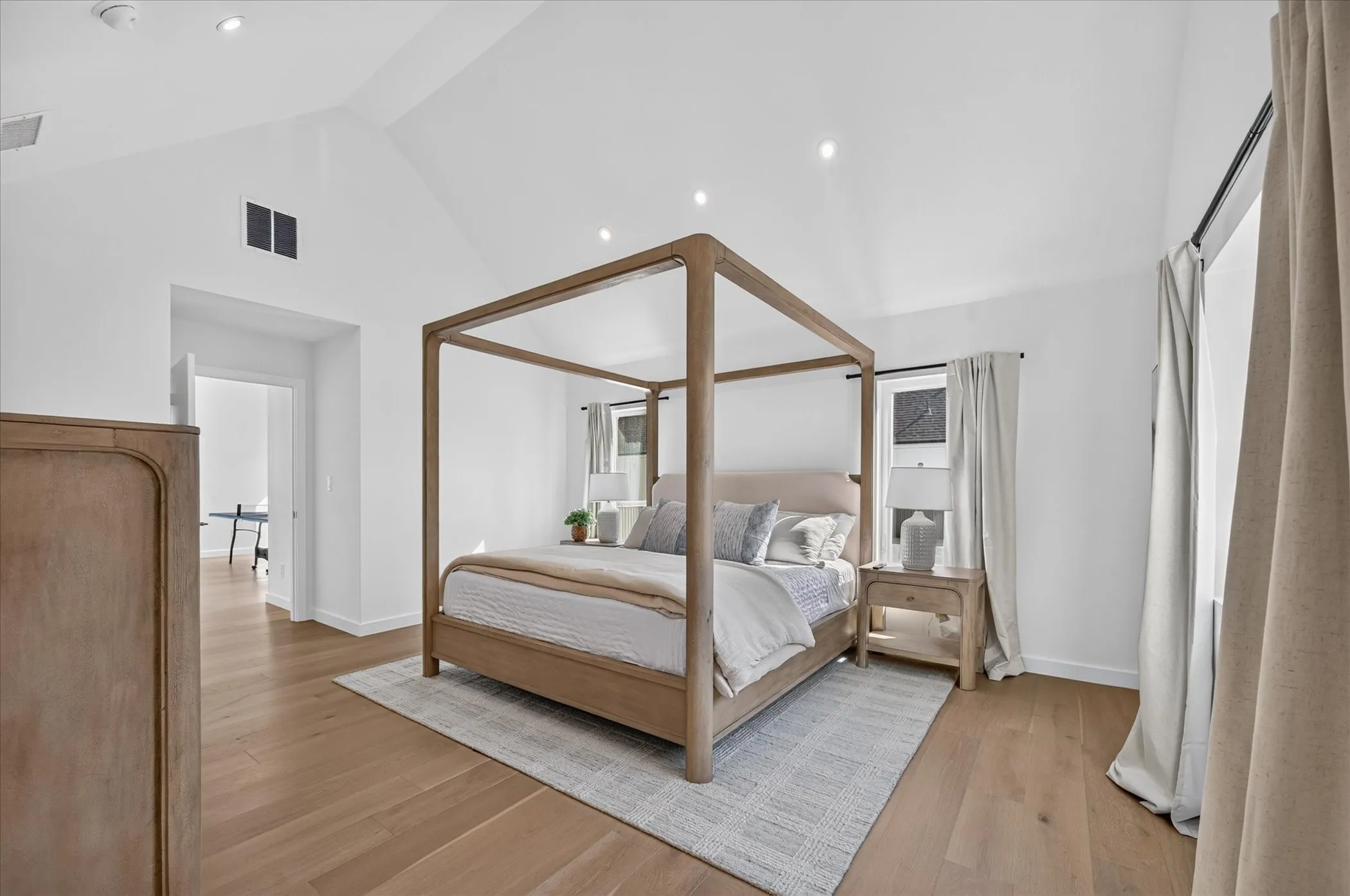 Bright primary bedroom showcasing a four-poster canopy bed frame in natural wood, wide-plank wood-finish flooring throughout, recessed ceiling lighting, and a neutral color palette with soft furnishings