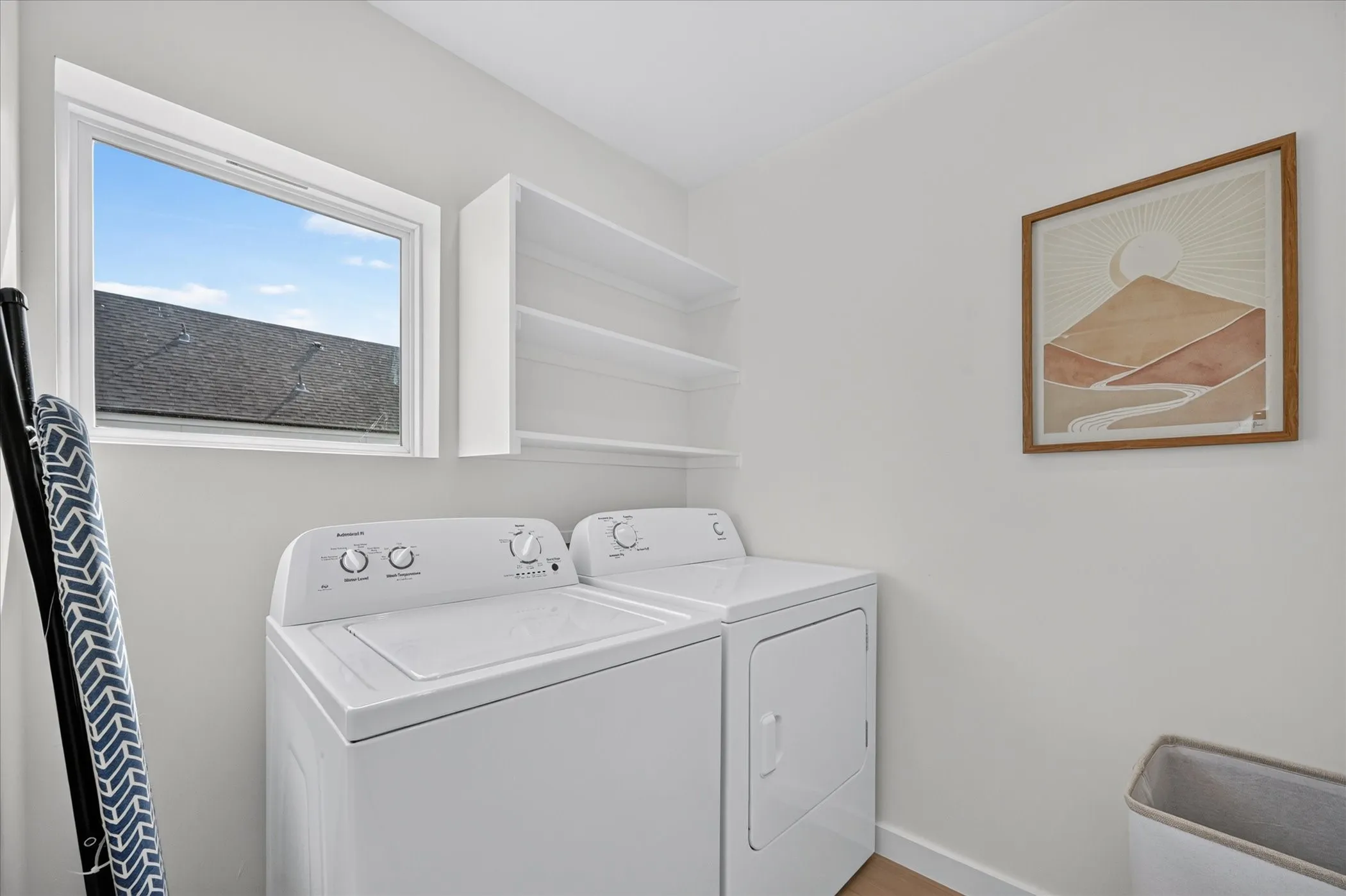 Dedicated laundry room featuring side-by-side washer and dryer with control-top design, white cabinetry with open shelving for storage, wood-finish flooring, and a transom window providing natural light