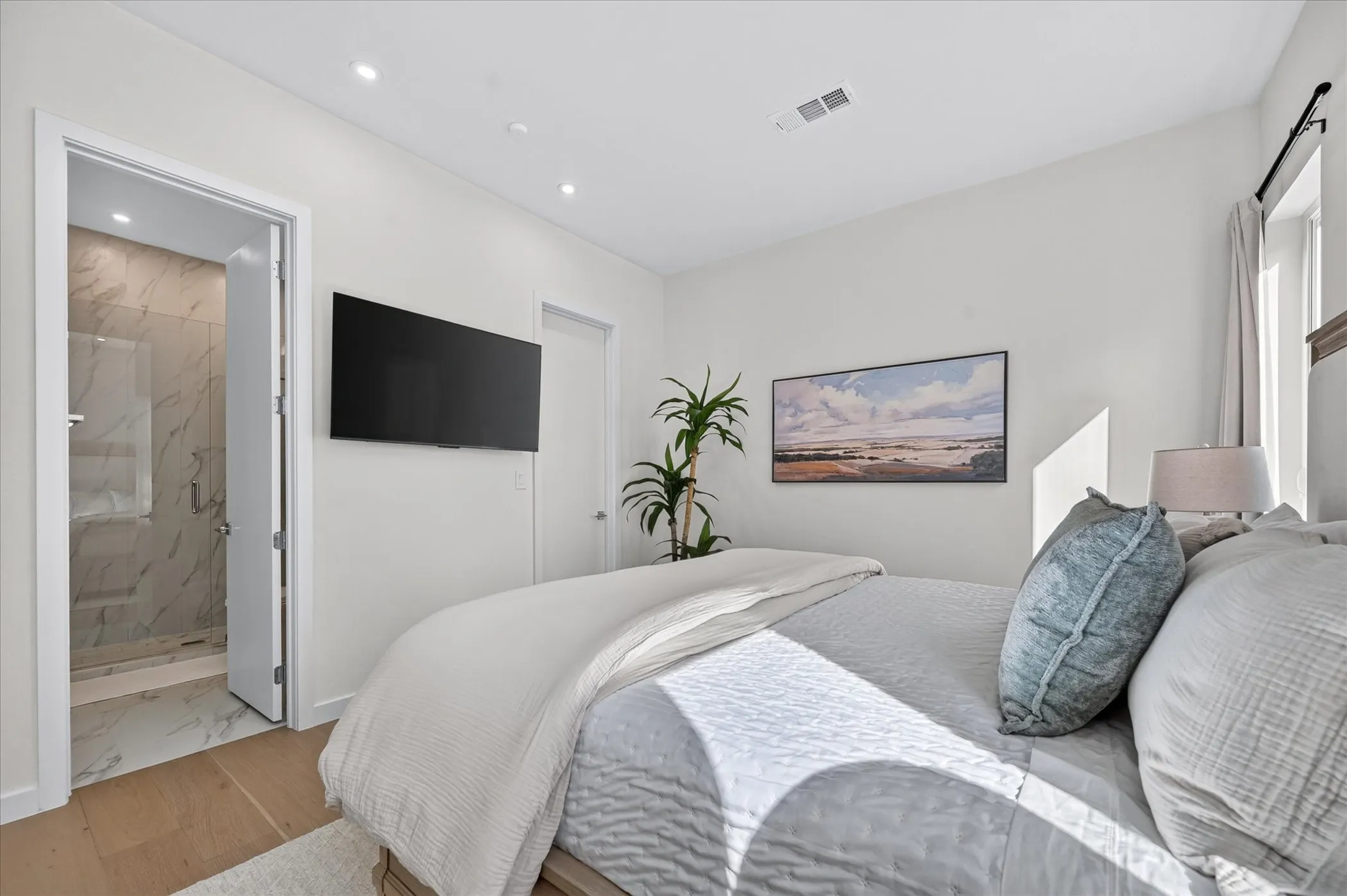 Light wood-finish flooring, recessed lighting, wall-mounted television, and a bathroom entry with full tile walls and glass shower enclosure
