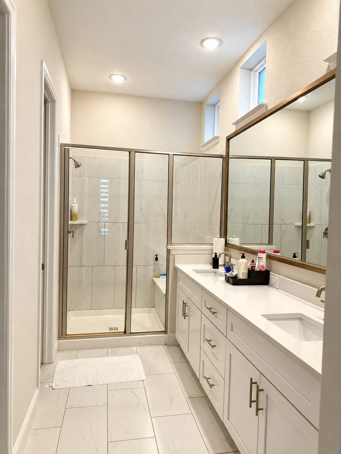 Spa-inspired primary bathroom featuring a frameless glass-enclosed shower with tile surround, dual vanity with white cabinetry and quartz countertop, large mirror with integrated lighting, recessed ceiling lights, and tile flooring throughout