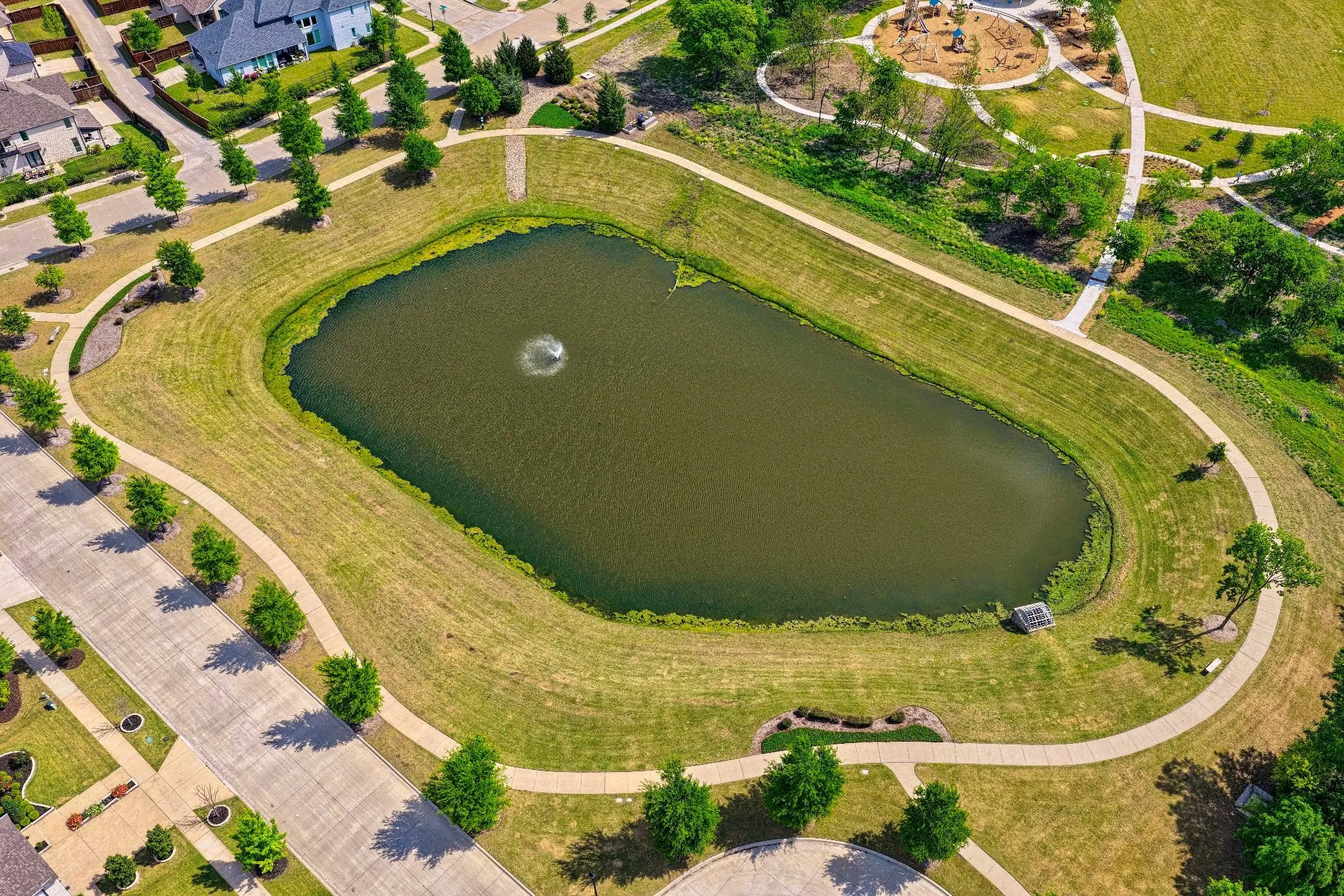 Aerial perspective featuring a pond with a water fountain, surrounded by manicured lawns and paved walking paths