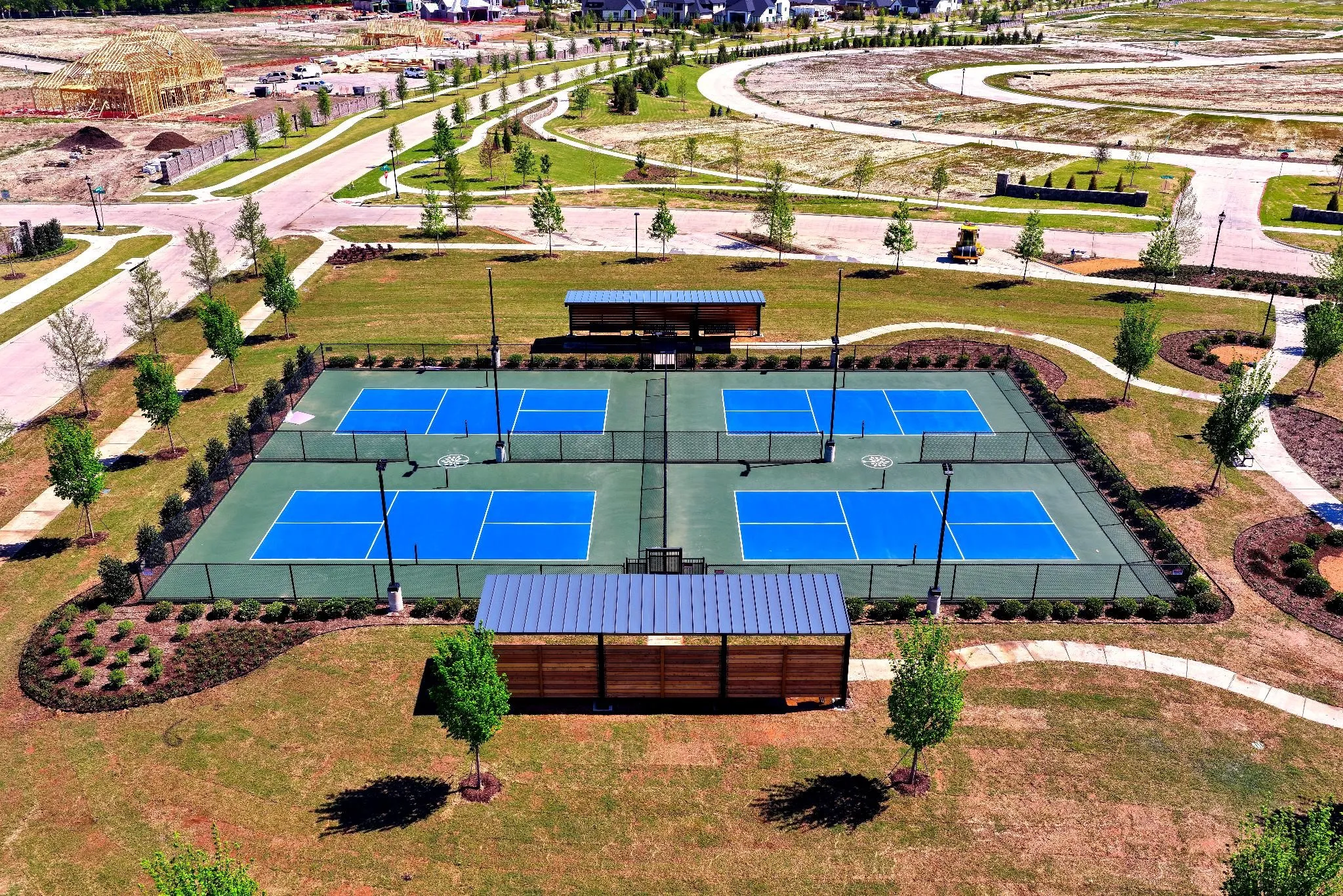 Four dedicated courts with blue playing surfaces, two covered seating areas, surrounding green landscaping, and paved walking paths