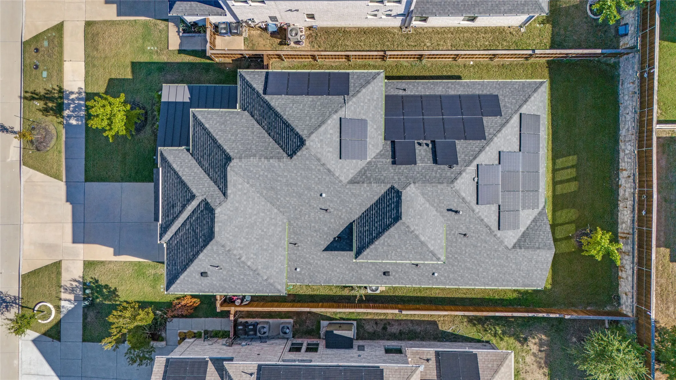 Gabled roofline with dark gray shingles, multiple solar panel installations, concrete driveway, concrete sidewalks, and a fenced perimeter