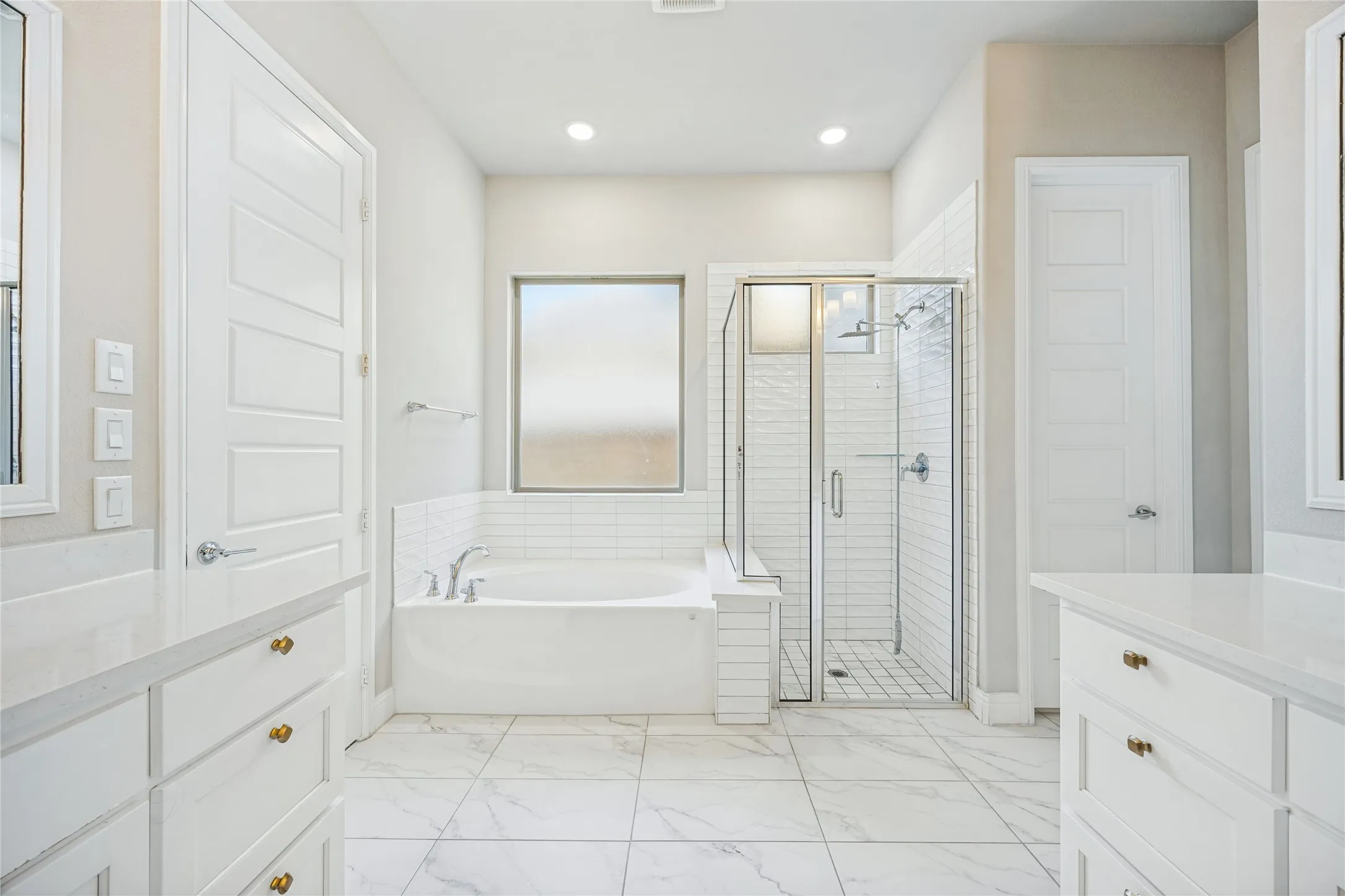 Spacious bathroom featuring a built-in bathtub, a walk-in shower with glass enclosure, dual vanities with white cabinetry, and large format floor tiling