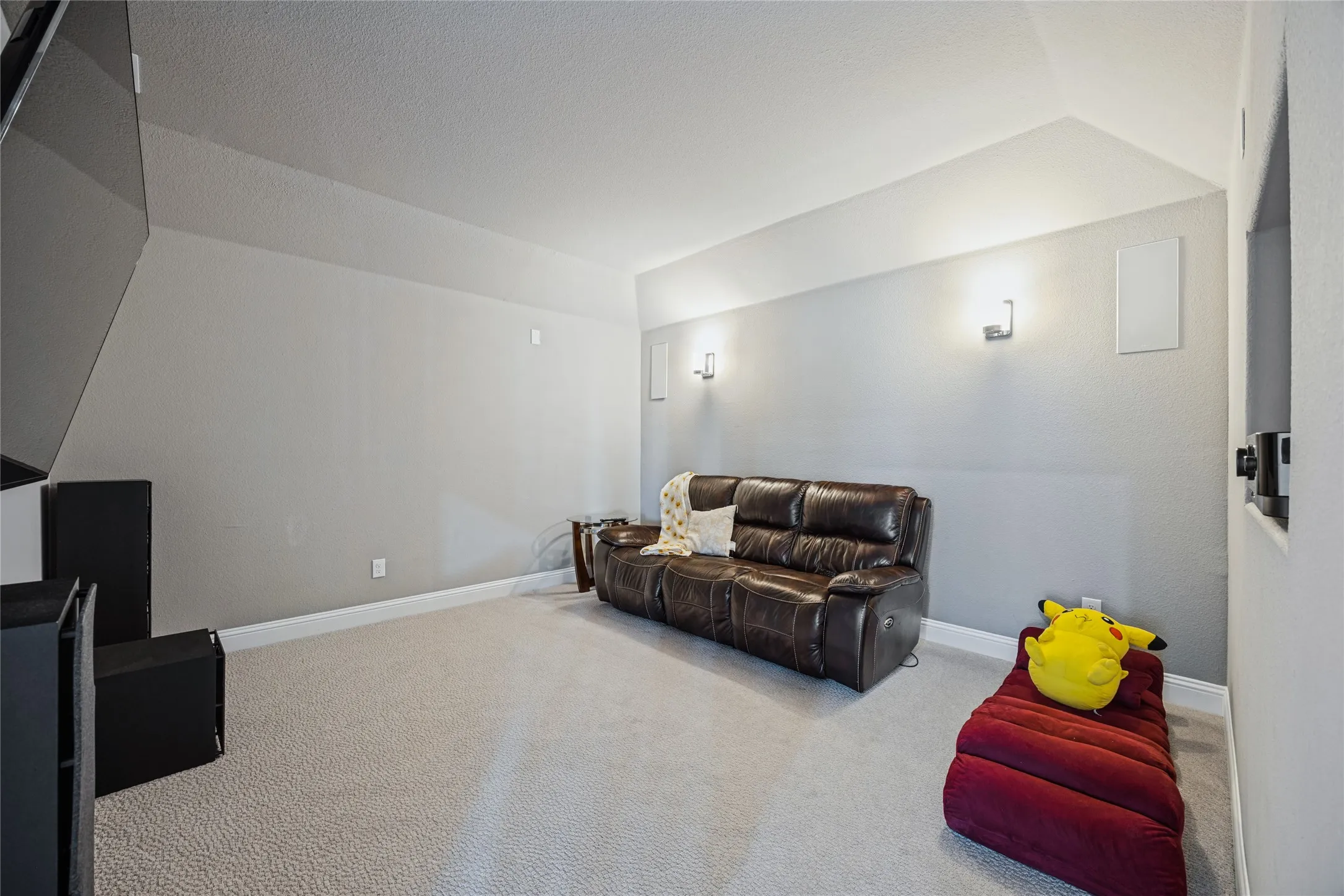Sloped ceiling with recessed lighting, wall-mounted light fixtures, textured carpet flooring, and integrated wall speakers