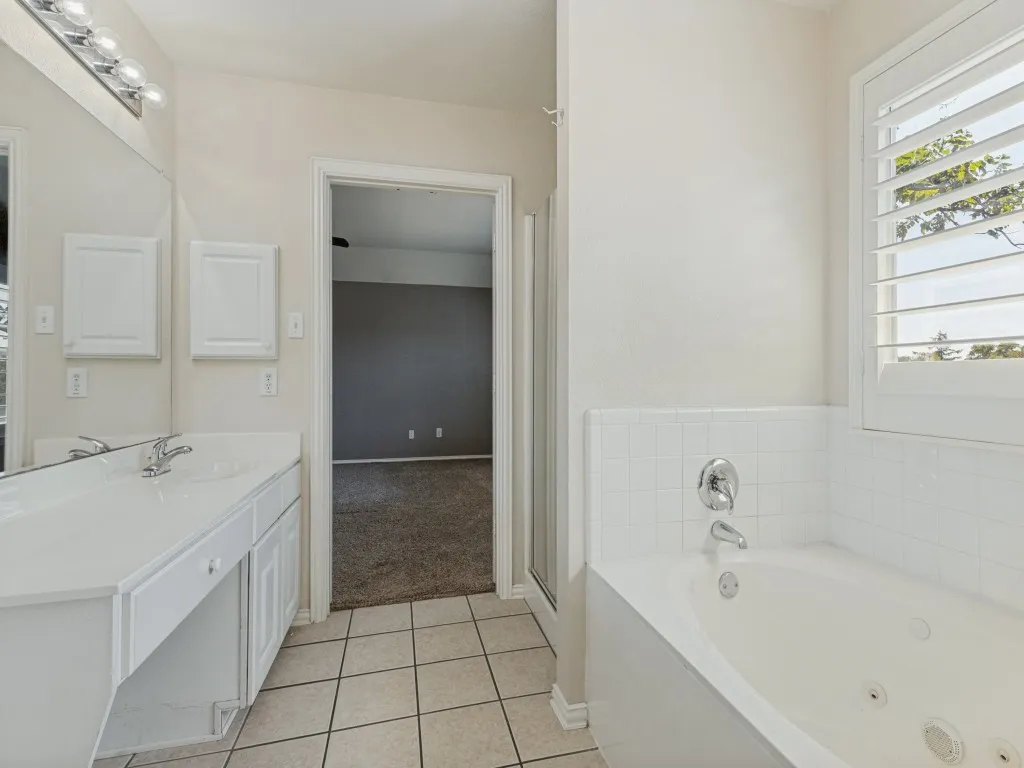 White tiled flooring, dual vanity with integrated sinks, recessed medicine cabinets, jetted soaking tub, and a window with plantation shutters