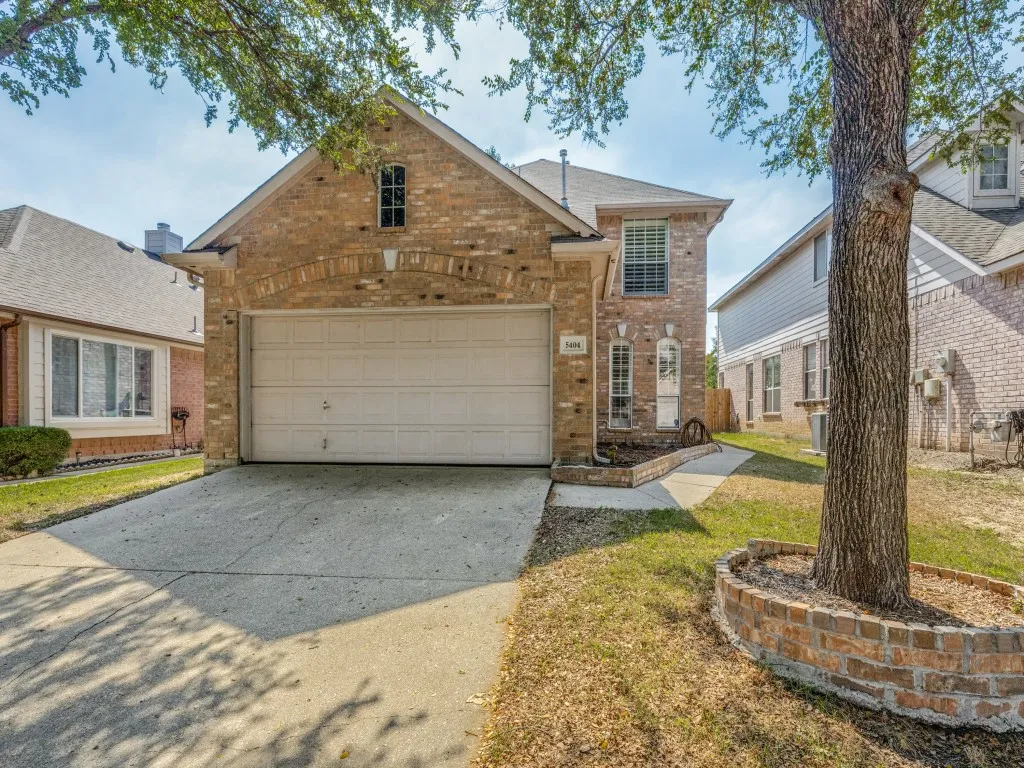Brick exterior with arched garage entry, concrete driveway, and mature tree with brick tree ring