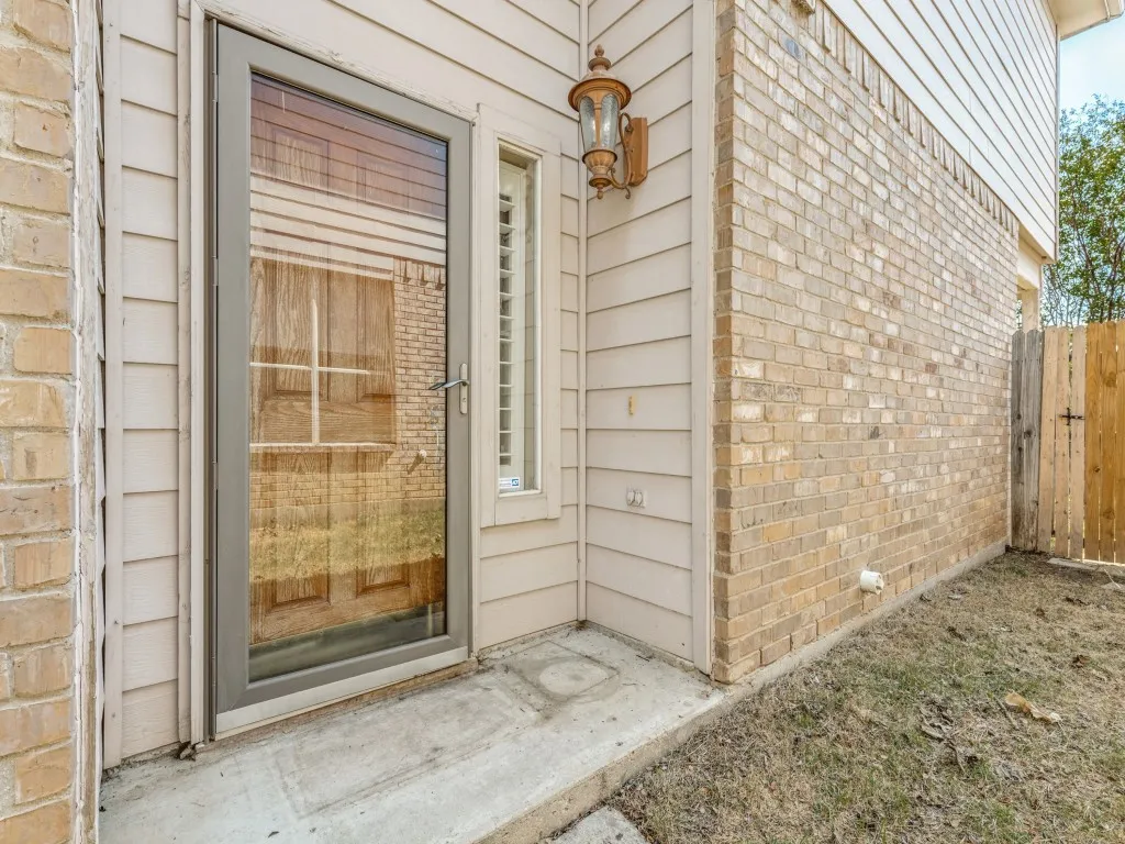 Full-light glass storm door, exterior wall lantern, brick and siding facade, concrete patio, and wood fence