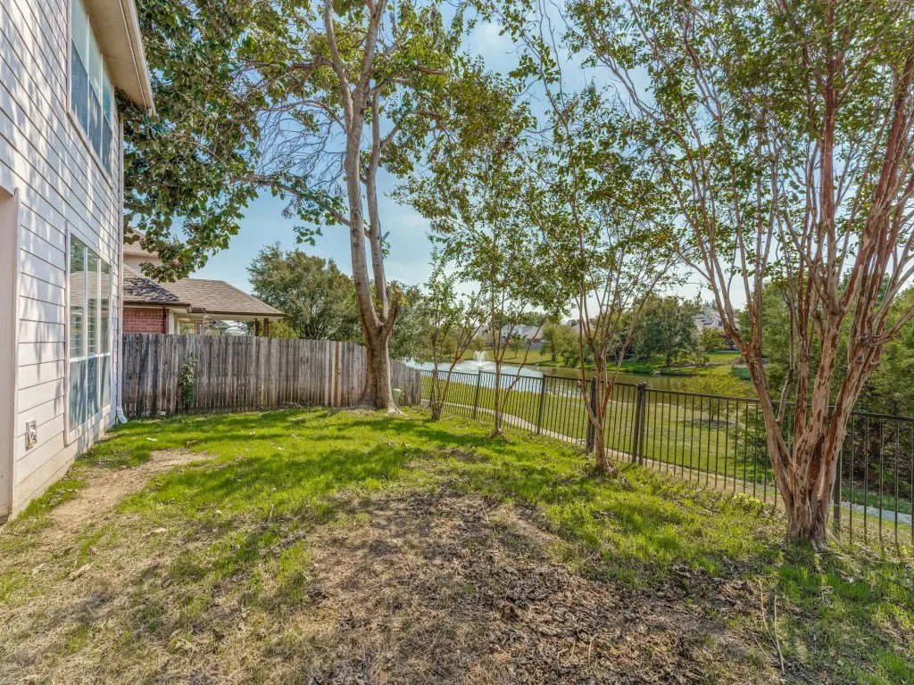 Expansive yard with mature trees, a wood privacy fence, and a black metal fence bordering a body of water with a water feature
