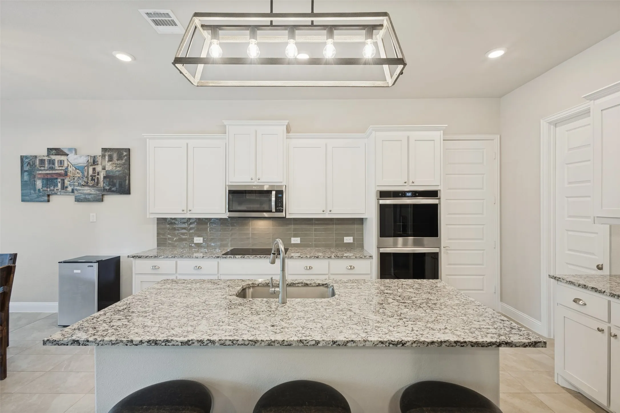 Kitchen featuring an island with stone countertop and undermount sink, white cabinetry with silver hardware, stainless steel built-in double oven, and gray tile backsplash