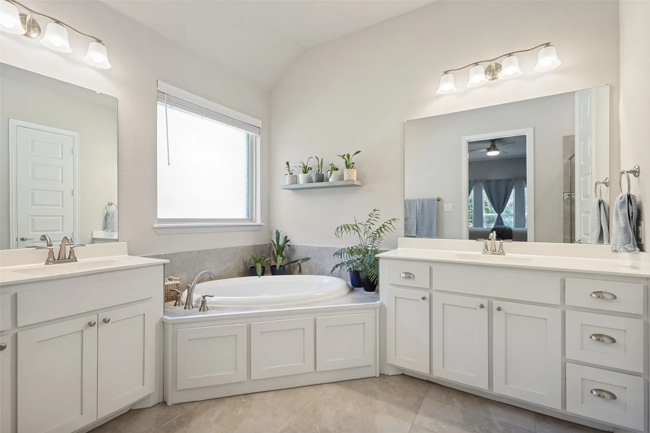 Dual vanity sinks with white cabinetry, a built-in soaking tub, a window with privacy blinds, and tile flooring