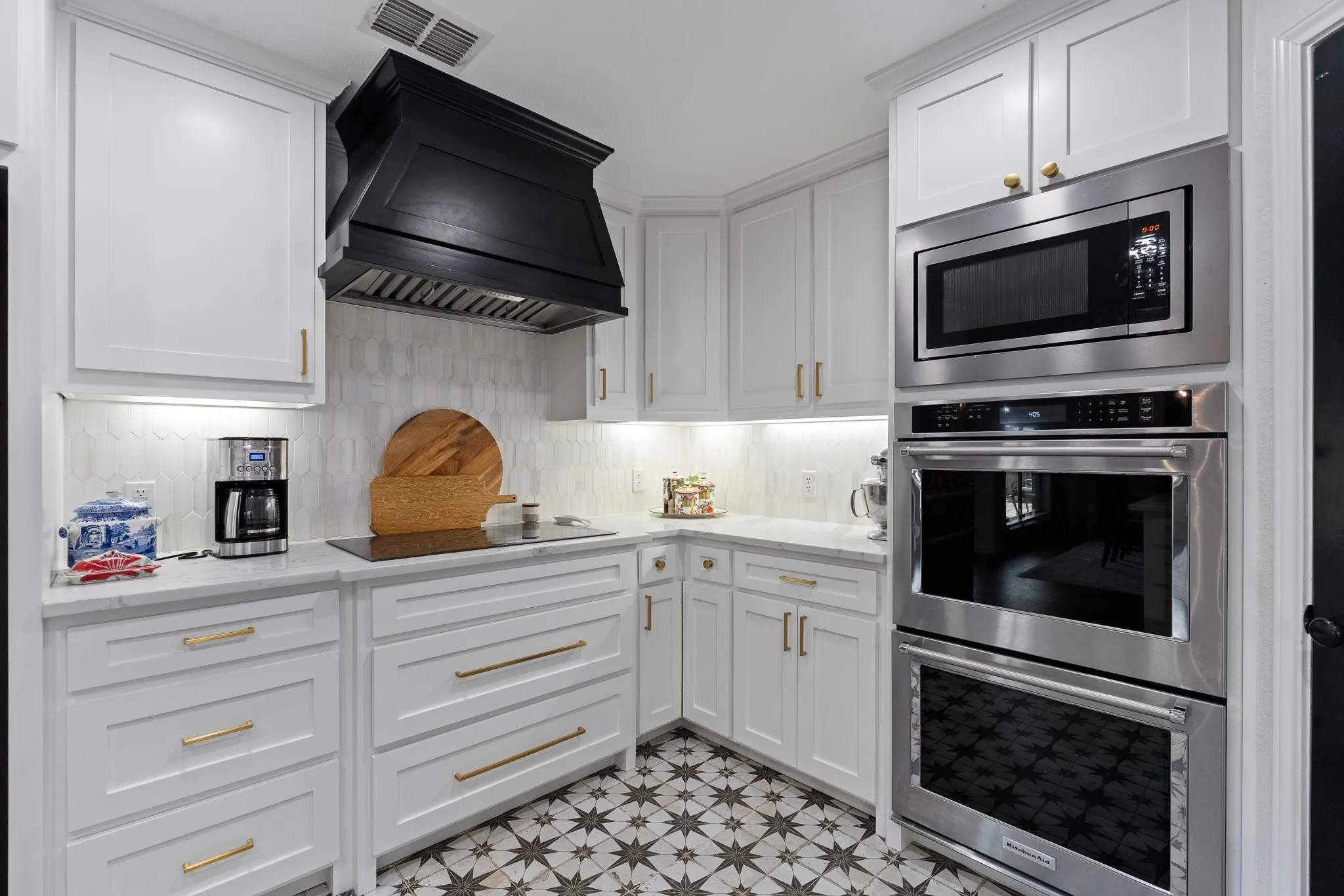 Modern kitchen featuring white cabinetry, light-colored countertops, a black range hood, stainless steel appliances, and designer floor tile