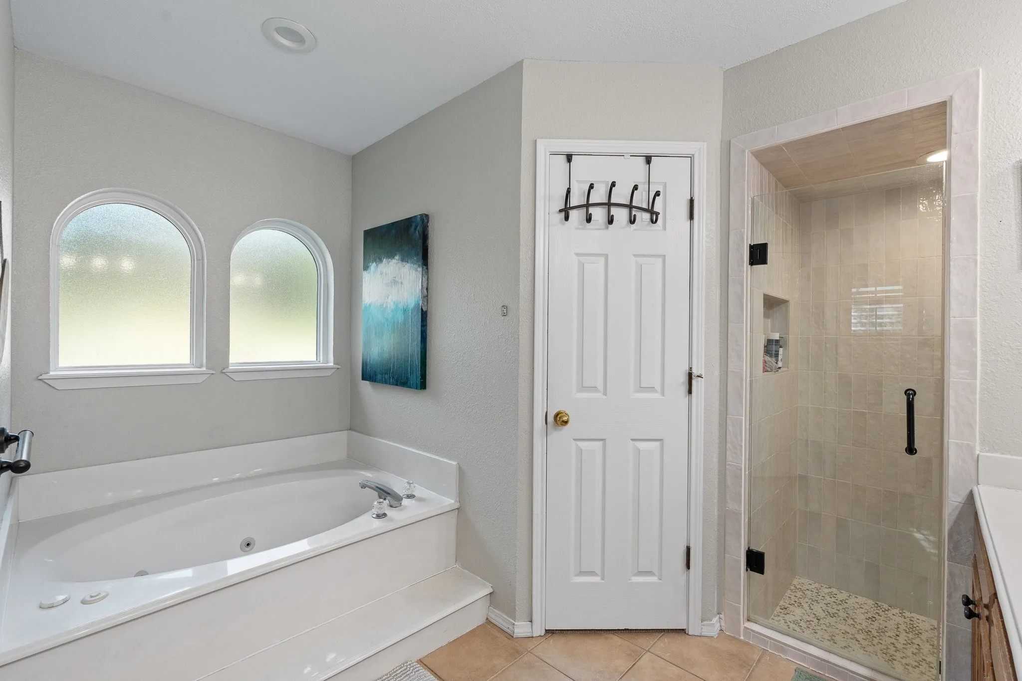 Jetted soaking tub with dual arched windows, new glass-enclosed shower with tile surround, and neutral tile flooring