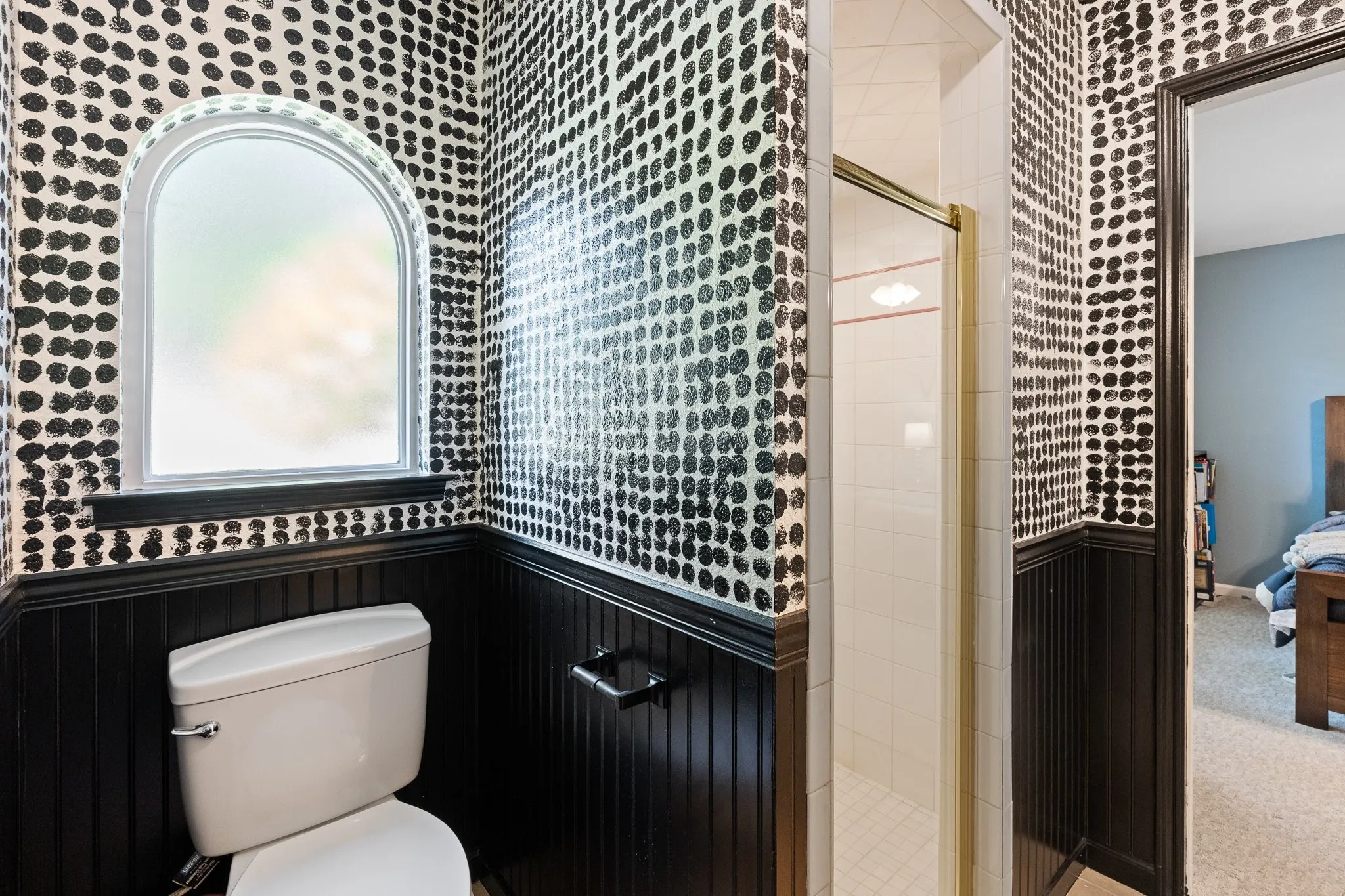 Arched window with privacy glass, black beadboard wainscoting, white toilet, and enclosed shower