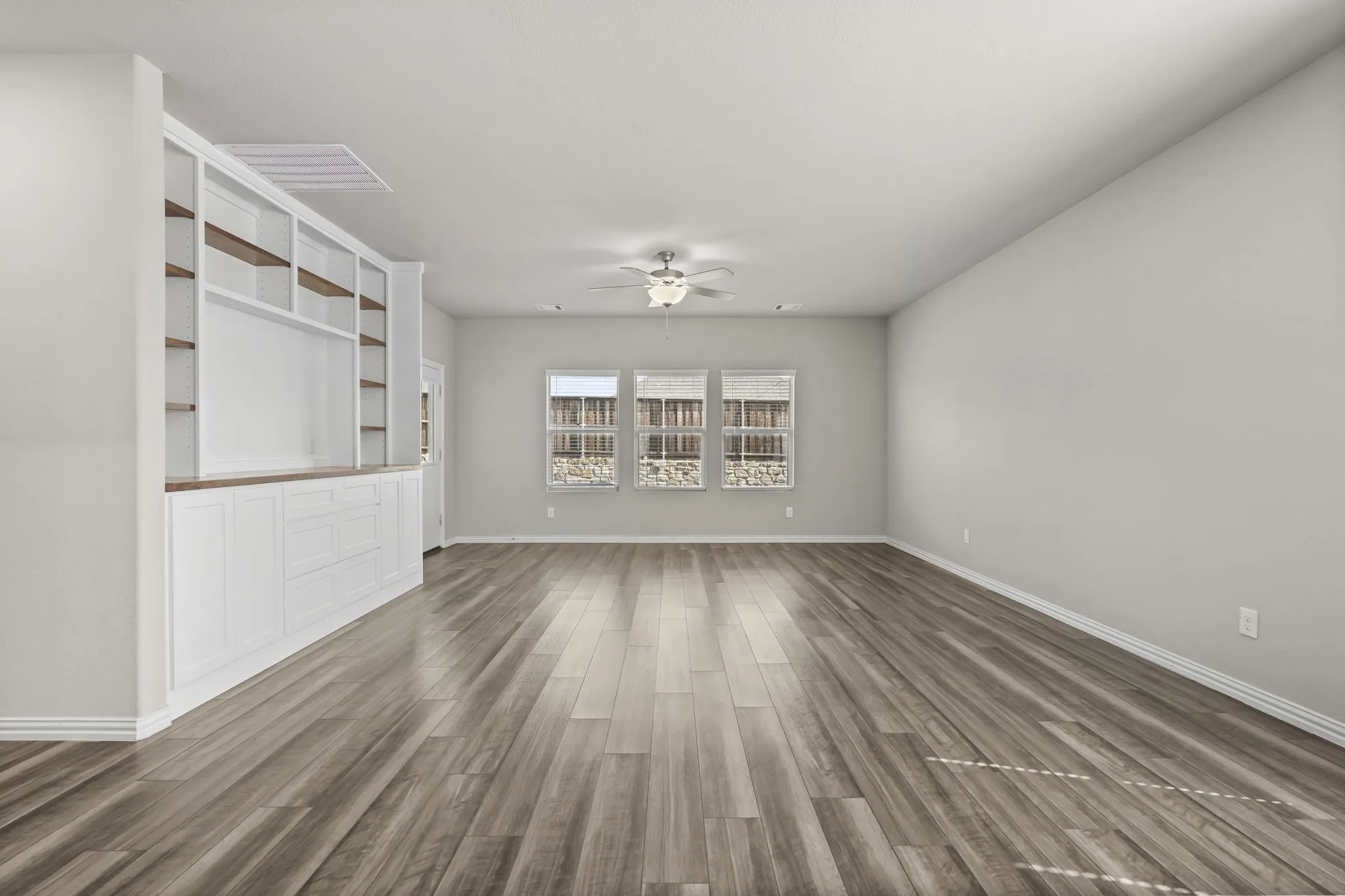 Spacious room featuring wood-finish flooring, three windows with blinds, a ceiling fan with integrated lighting, and a built-in white media center with open shelving and lower cabinetry
