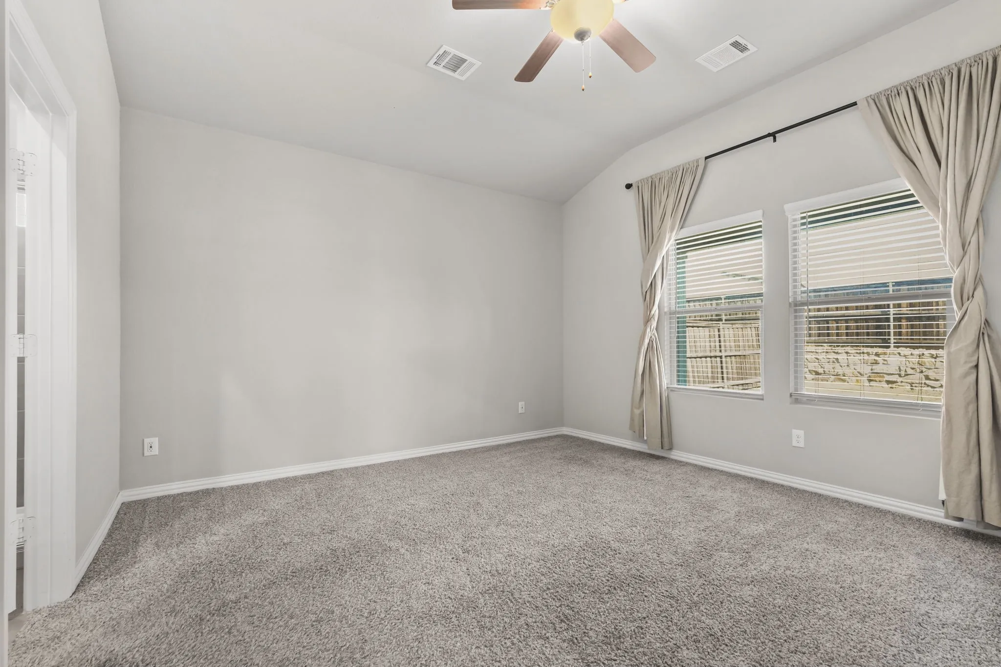 Vaulted ceiling, two windows with blinds, ceiling fan with light, light grey carpeting, and white baseboards