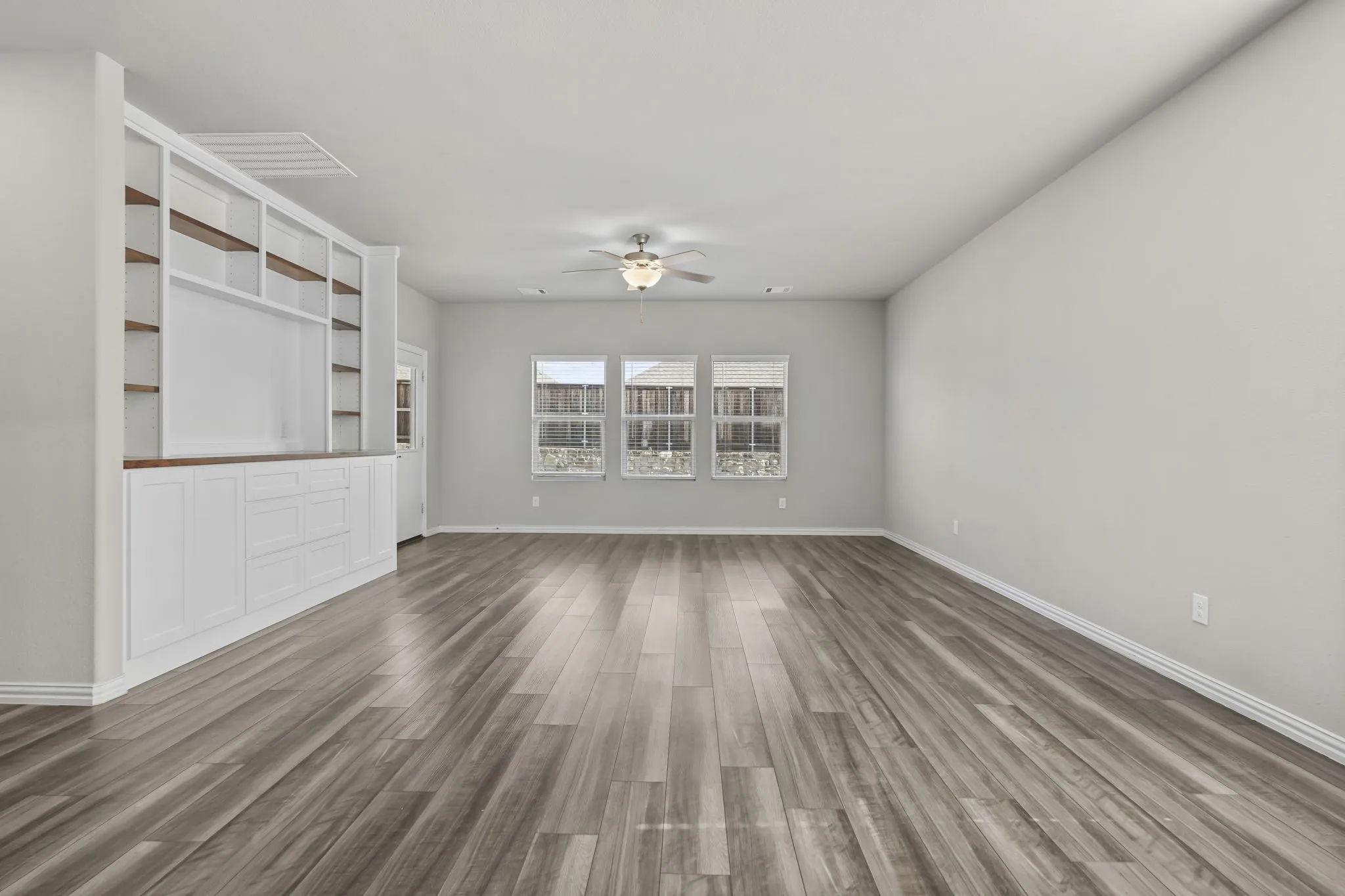 Expansive room featuring wood-finish flooring, three windows with white blinds, a ceiling fan with integrated lighting, and extensive built-in cabinetry with open shelving and closed storage