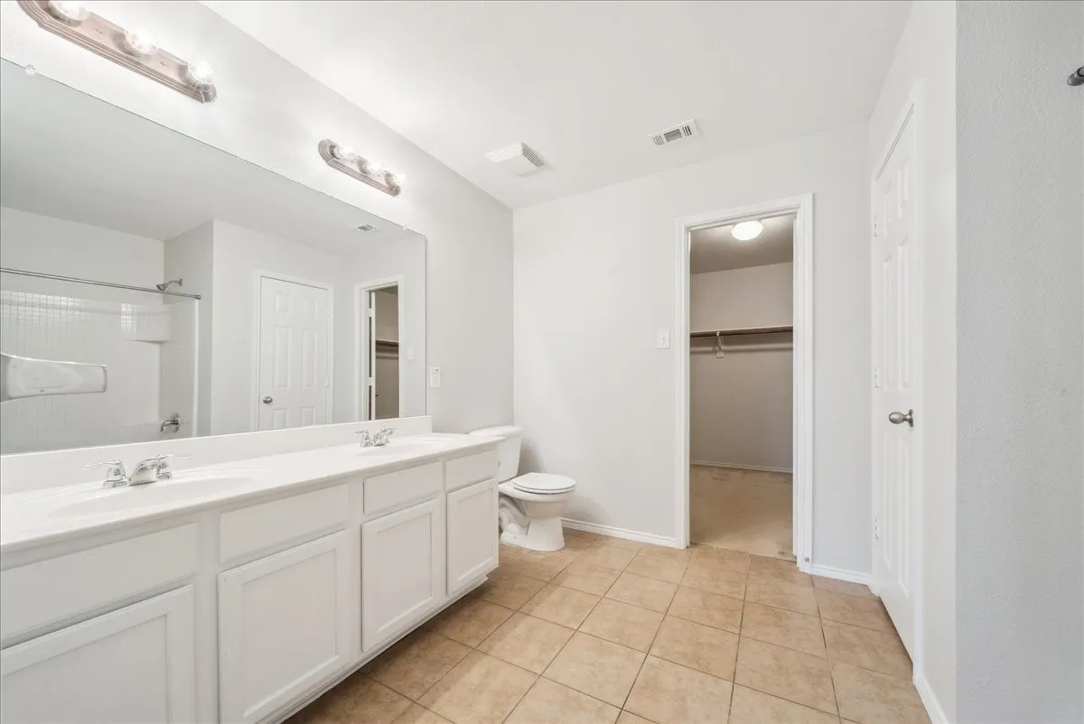 Dual vanity with white cabinetry, polished chrome fixtures, expansive mirror, tiled flooring, and a walk-in closet with shelving