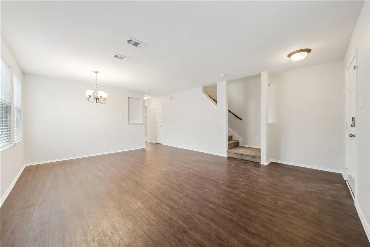 Wood-finish flooring throughout, bright white walls, multiple windows with horizontal blinds, overhead chandelier, and a carpeted staircase