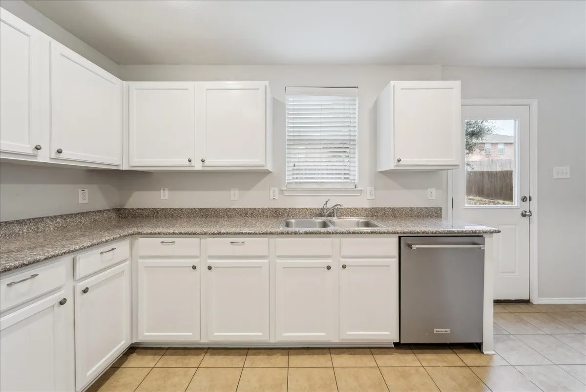 Kitchen featuring white shaker-style cabinetry, a stainless steel double basin sink, stone-look countertops, and a tile flooring