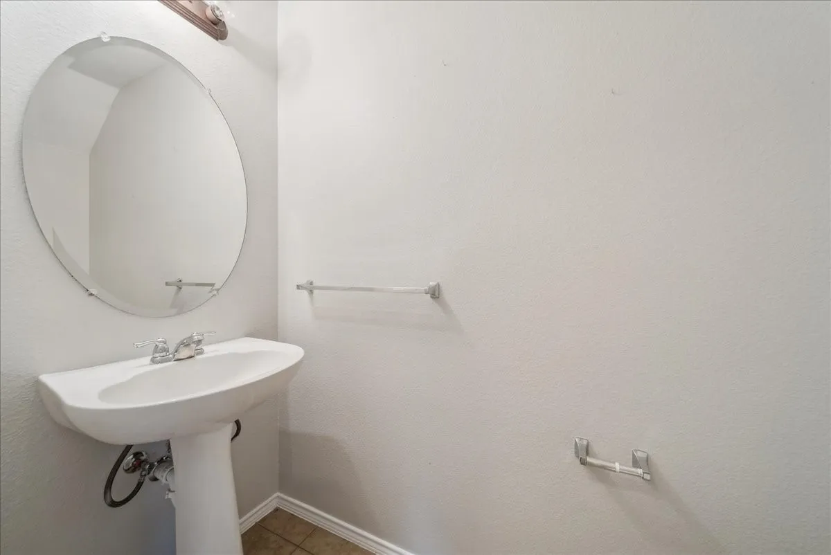 Pedestal sink with chrome fixtures and an oval mirror, complemented by tile flooring and a wall-mounted light fixture