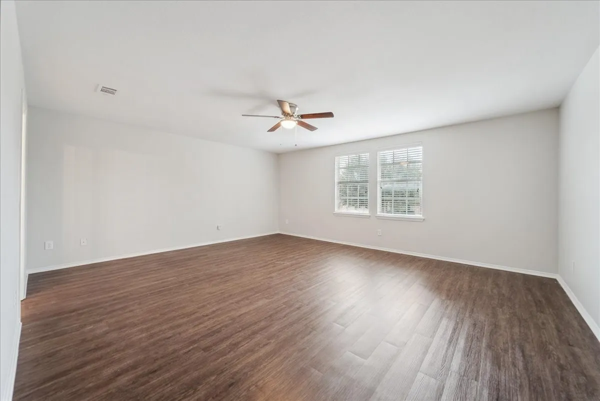 Spacious room featuring wood-finish flooring, white walls, dual windows with blinds, and a ceiling fan