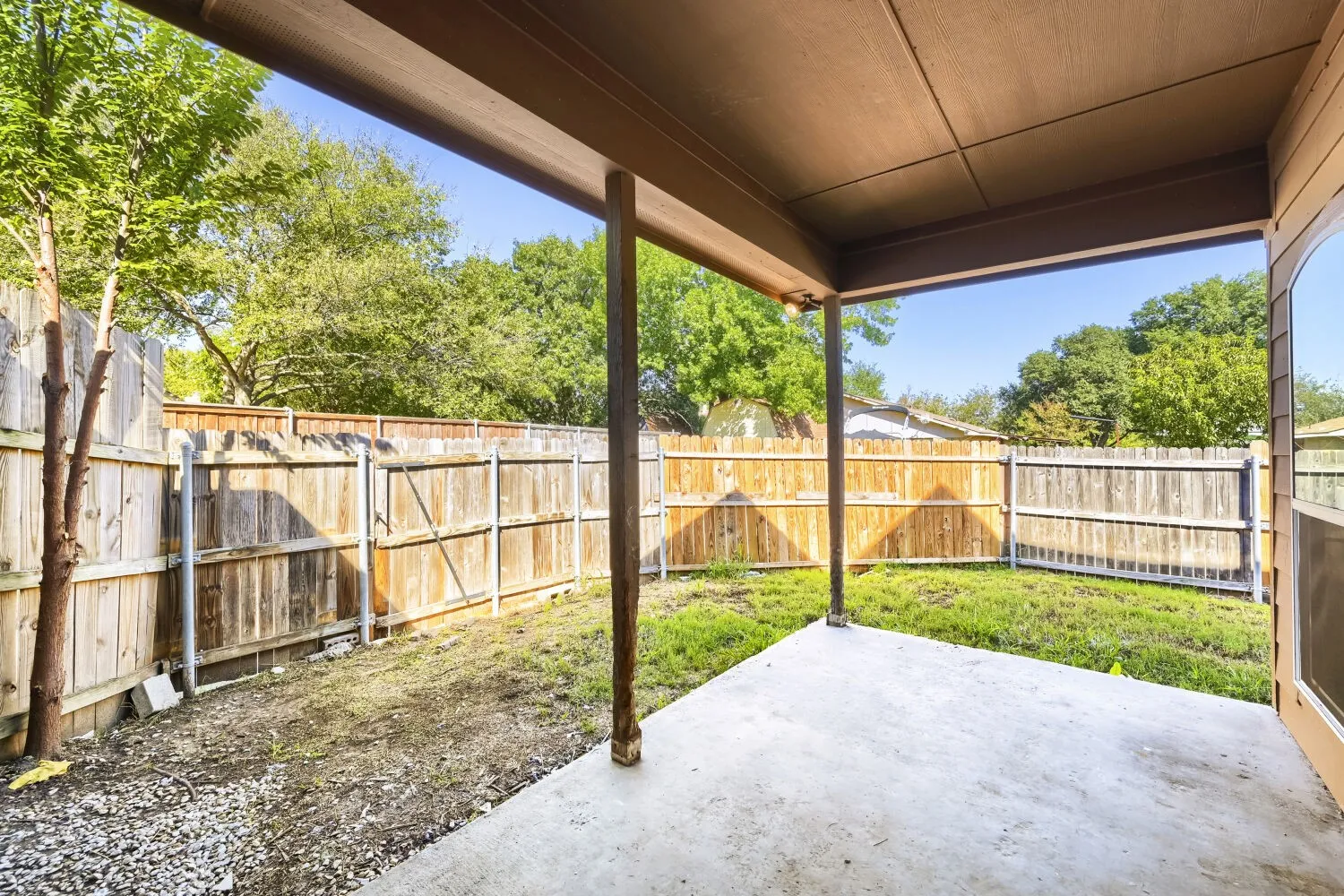 Covered concrete patio with wooden support columns, enclosed by wood fencing and a grassy yard