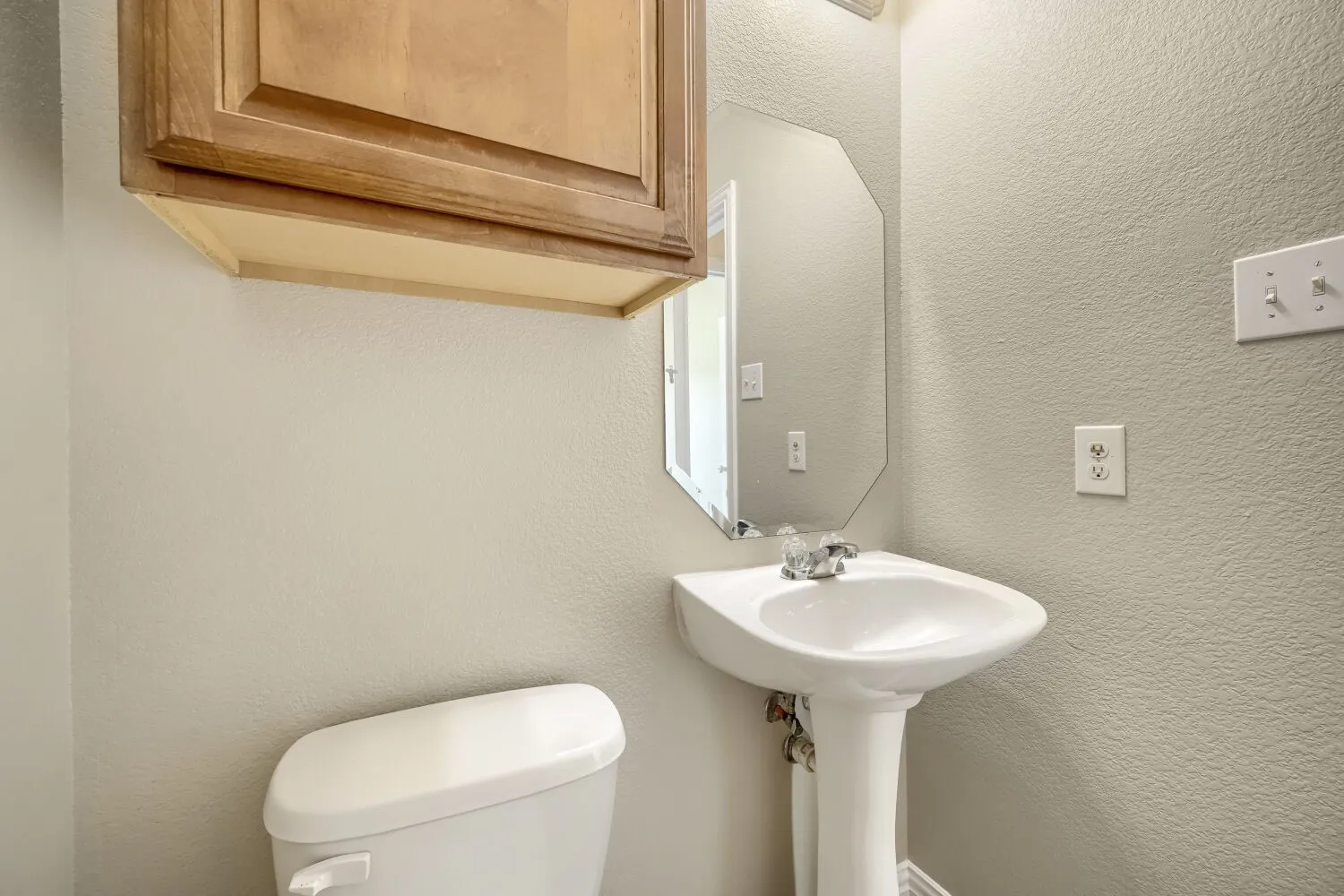 Pedestal sink with chrome faucet, octagonal wall mirror, light wood-finish cabinetry, and neutral wall tones