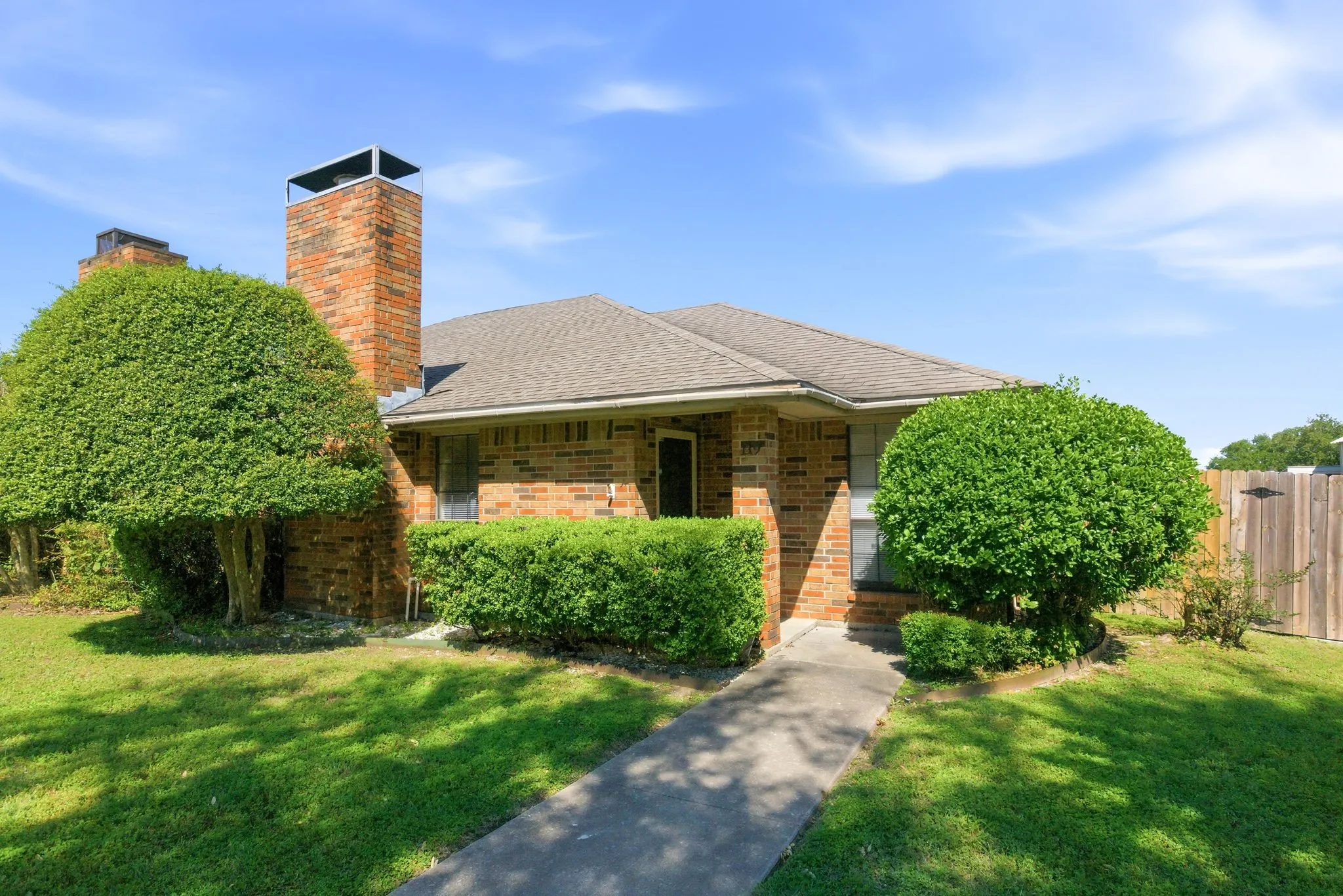 Brick exterior, twin brick chimneys, concrete walkway, pitched roof, and mature landscaping