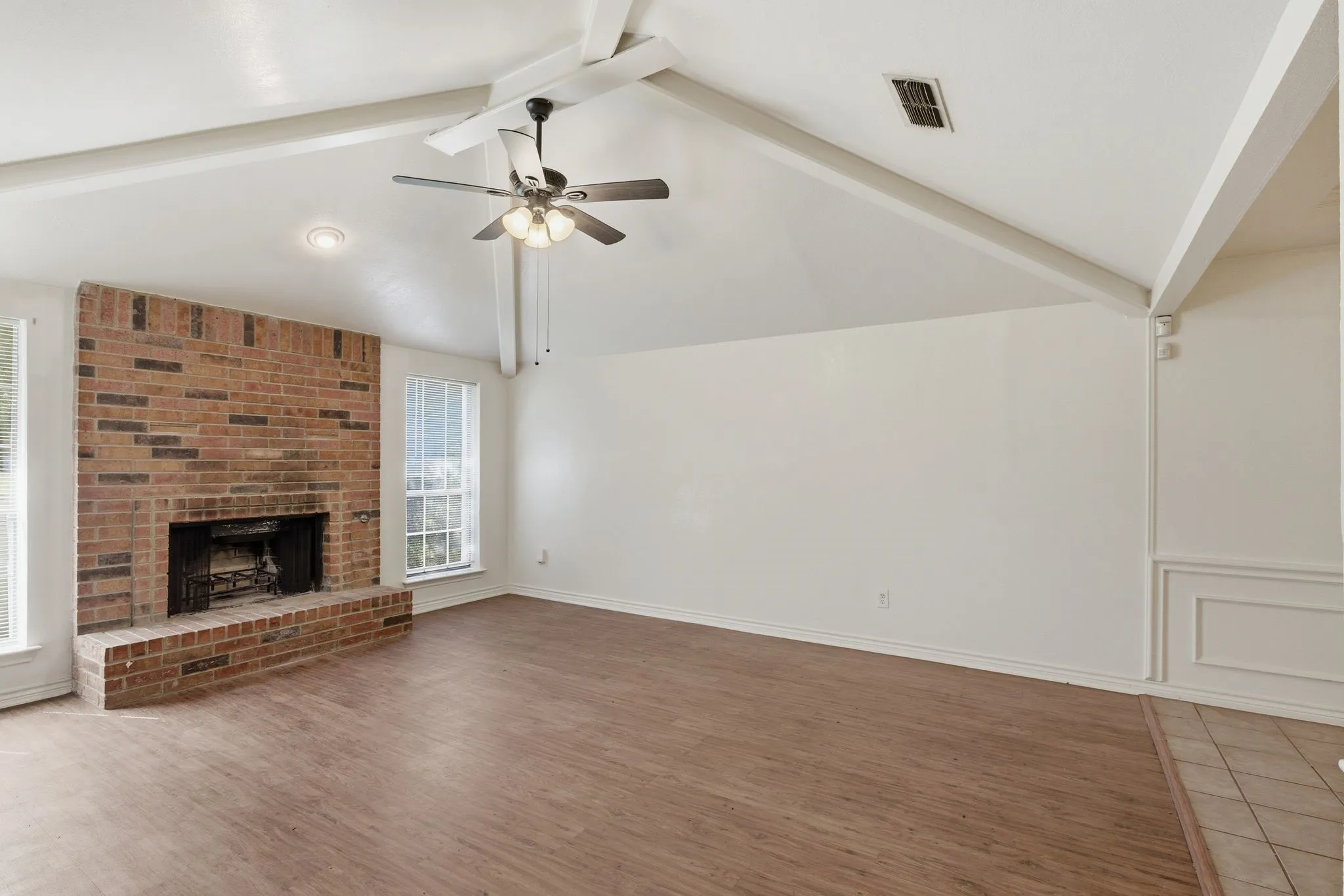 Vaulted ceiling with exposed beams, brick fireplace, wood-finish flooring, and a ceiling fan with integrated lighting