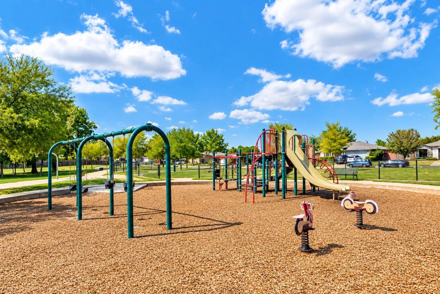 Recreational playground area featuring a swing set, multi-level play structure with slides, spring riders, and a wood chip surface