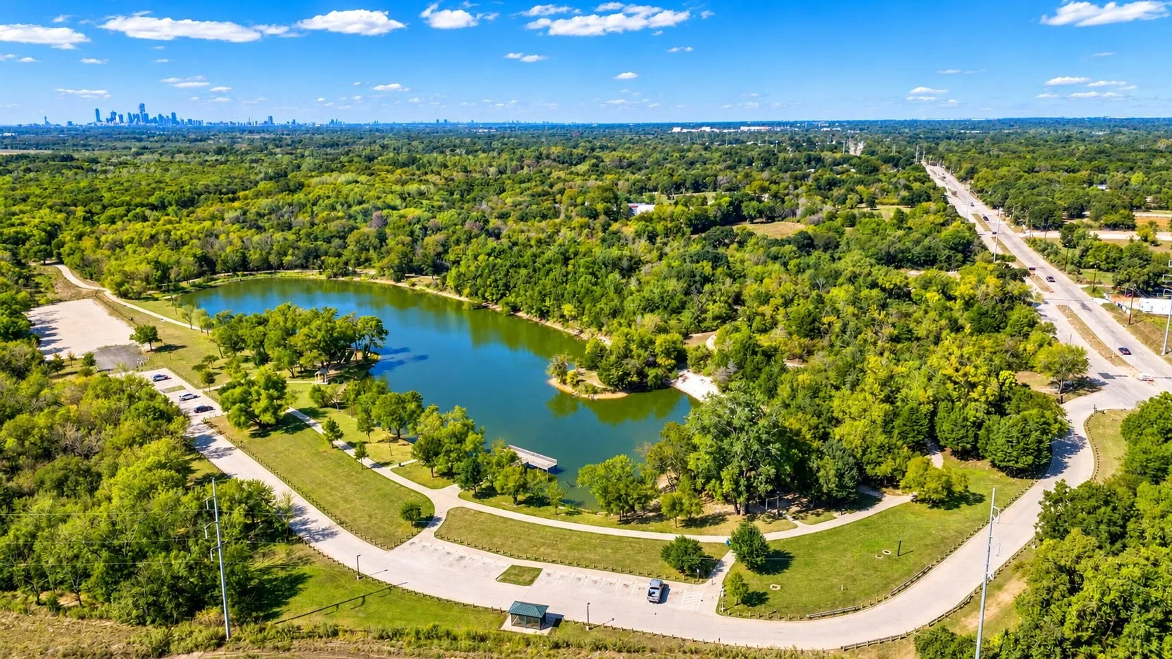 Lakeside property with a paved roadway, abundant trees, and distant city skyline