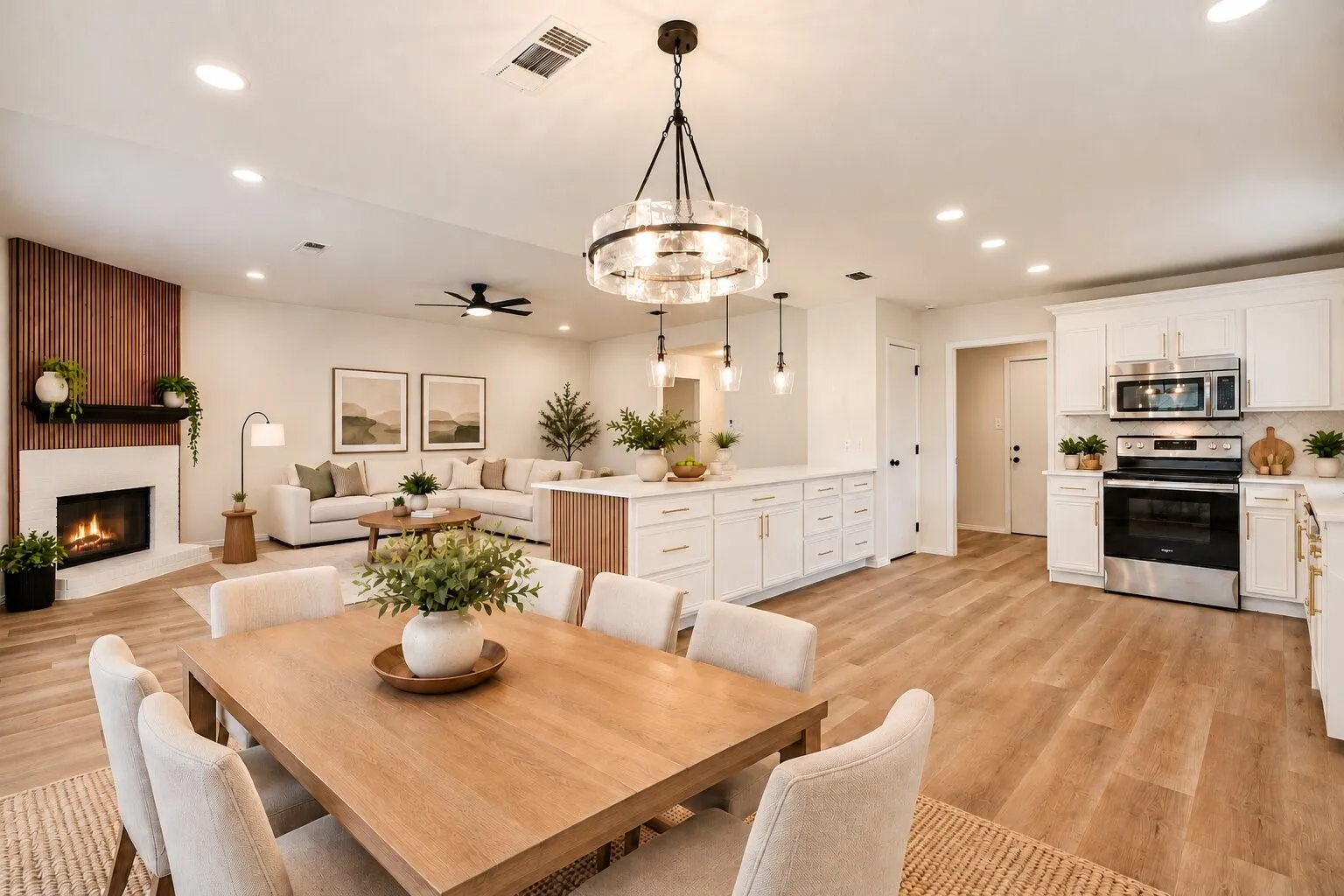 Open-concept living space featuring wood-finish flooring, a contemporary fireplace with a wood-paneled accent wall, a kitchen island with storage, and a dining area illuminated by a modern chandelier