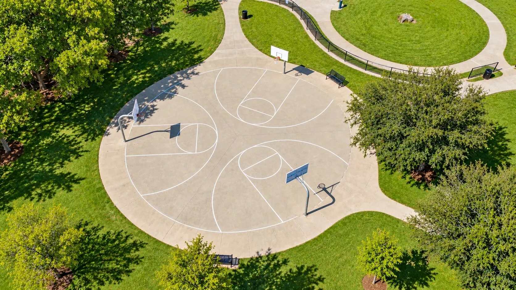 Concrete multi-court basketball area with three hoops, surrounded by green landscaping and walking paths