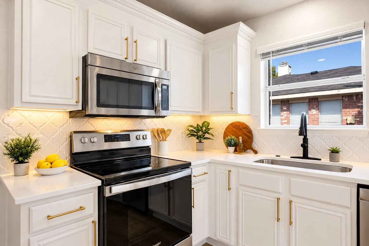 White cabinetry with brass-finish hardware, under-cabinet lighting, a window over the sink, a stainless steel sink, and white countertops
