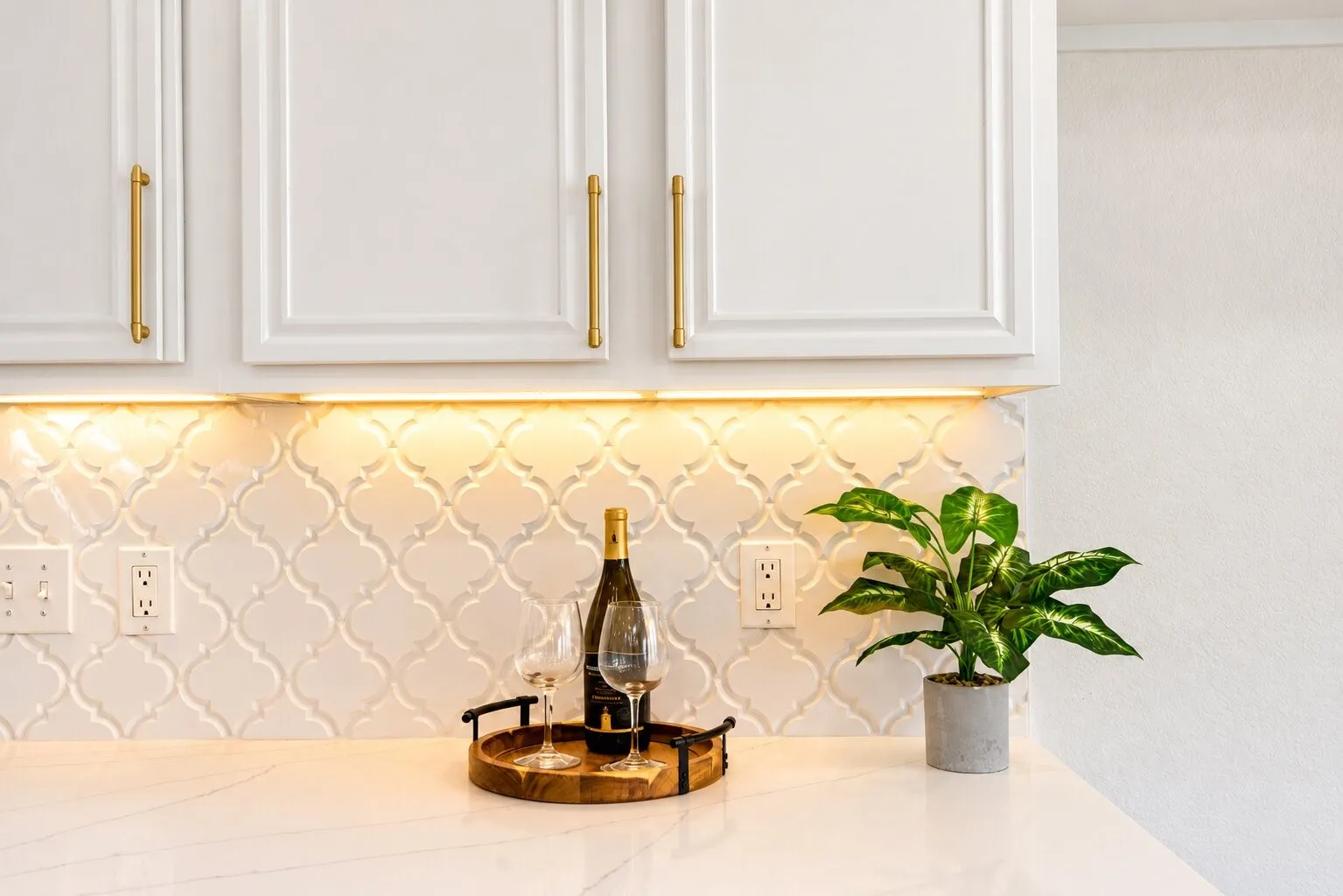 White cabinetry with gold-tone hardware, under-cabinet lighting, arabesque tile backsplash, and a solid surface countertop