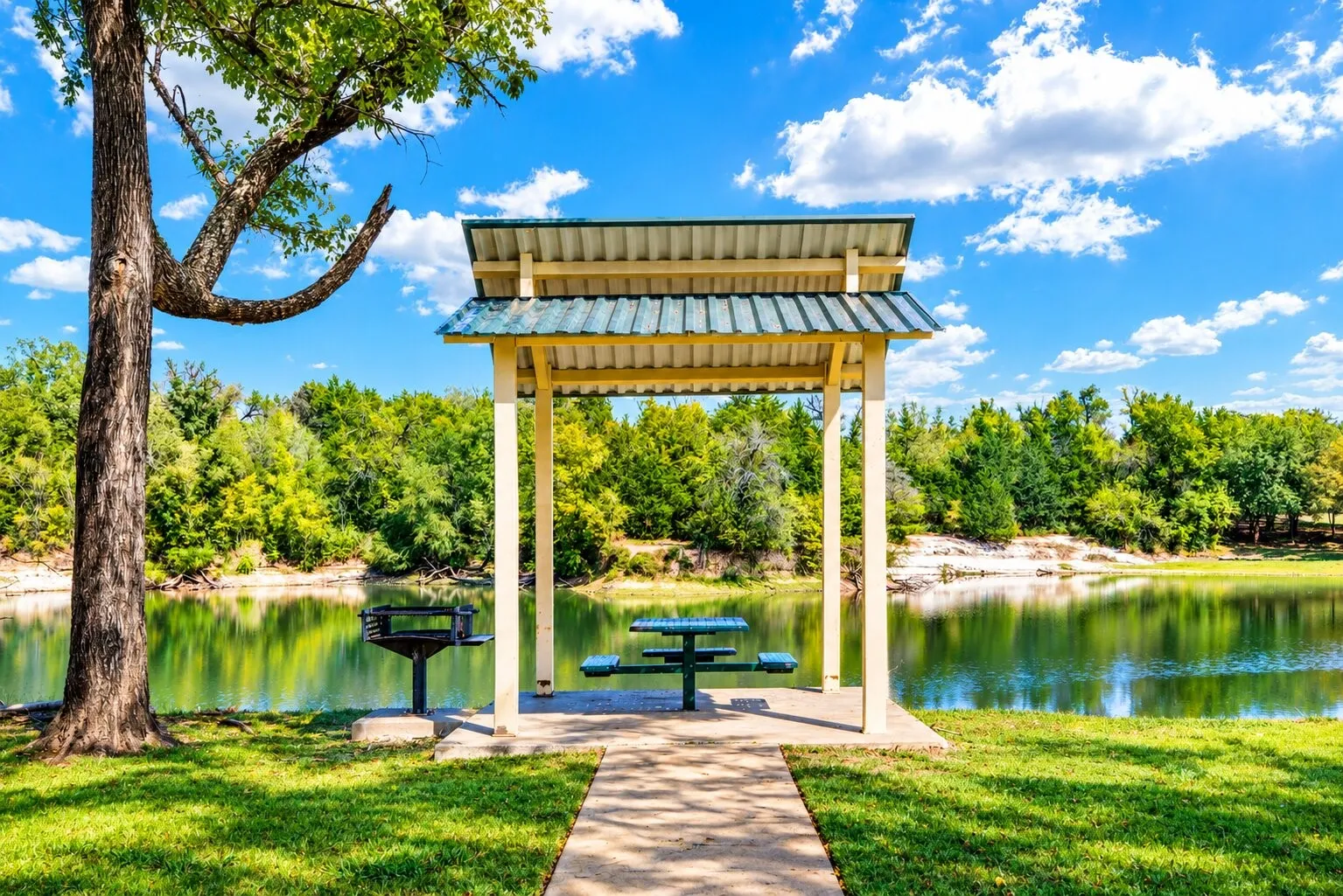 Lakeside pavilion with a built-in picnic table, adjacent to a charcoal grill and a concrete pathway