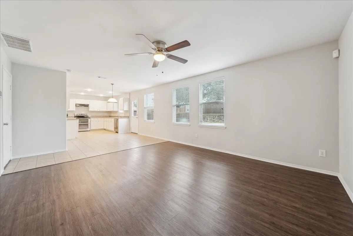 Wood-finish flooring, white walls, ceiling fan, multiple windows, and a partially visible kitchen with white cabinetry
