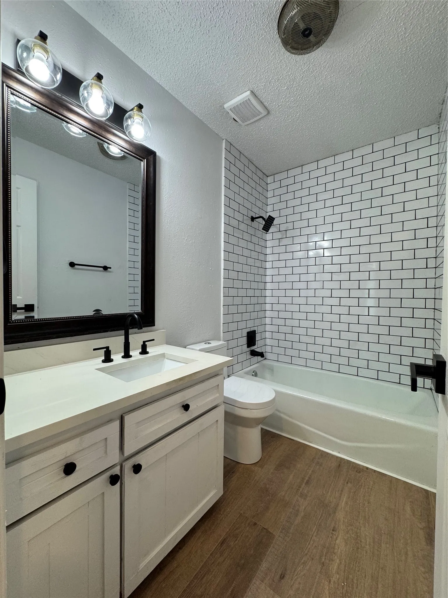 White subway tile shower surround, wood-finish flooring, white vanity with integrated sink, dark bronze hardware, and a framed mirror