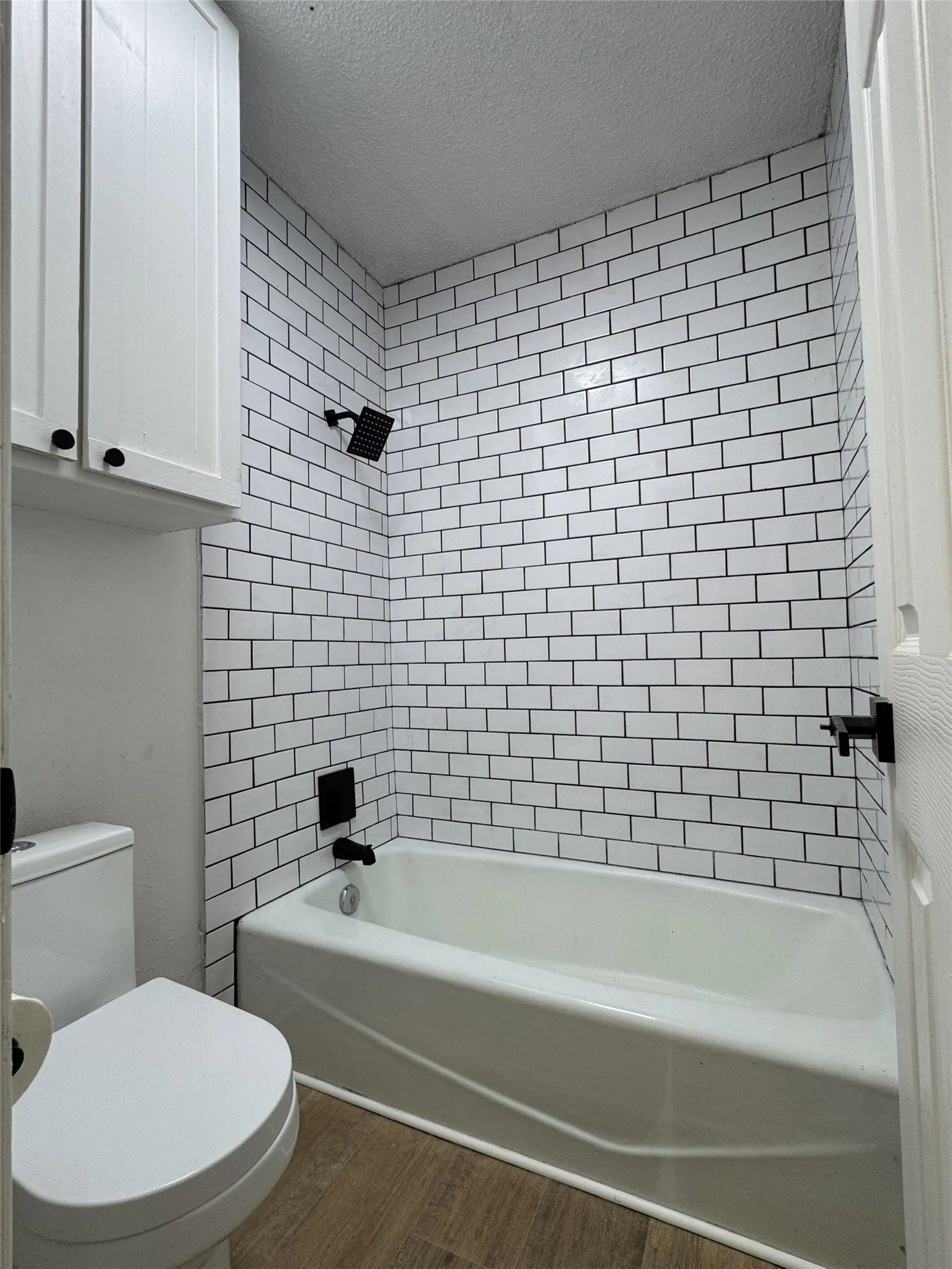 White subway tile surround with dark grout, matte black shower fixtures, white bathtub, wood-finish flooring, and white upper cabinetry