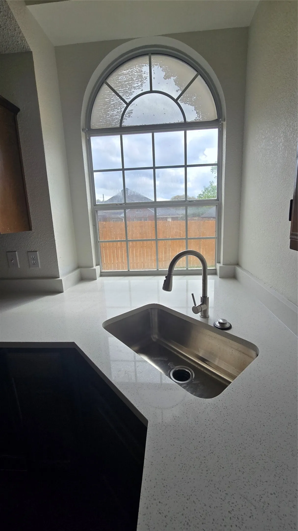 Arched window with divided lites, undermount stainless steel sink, gooseneck faucet, light-toned countertop, and dark base cabinetry