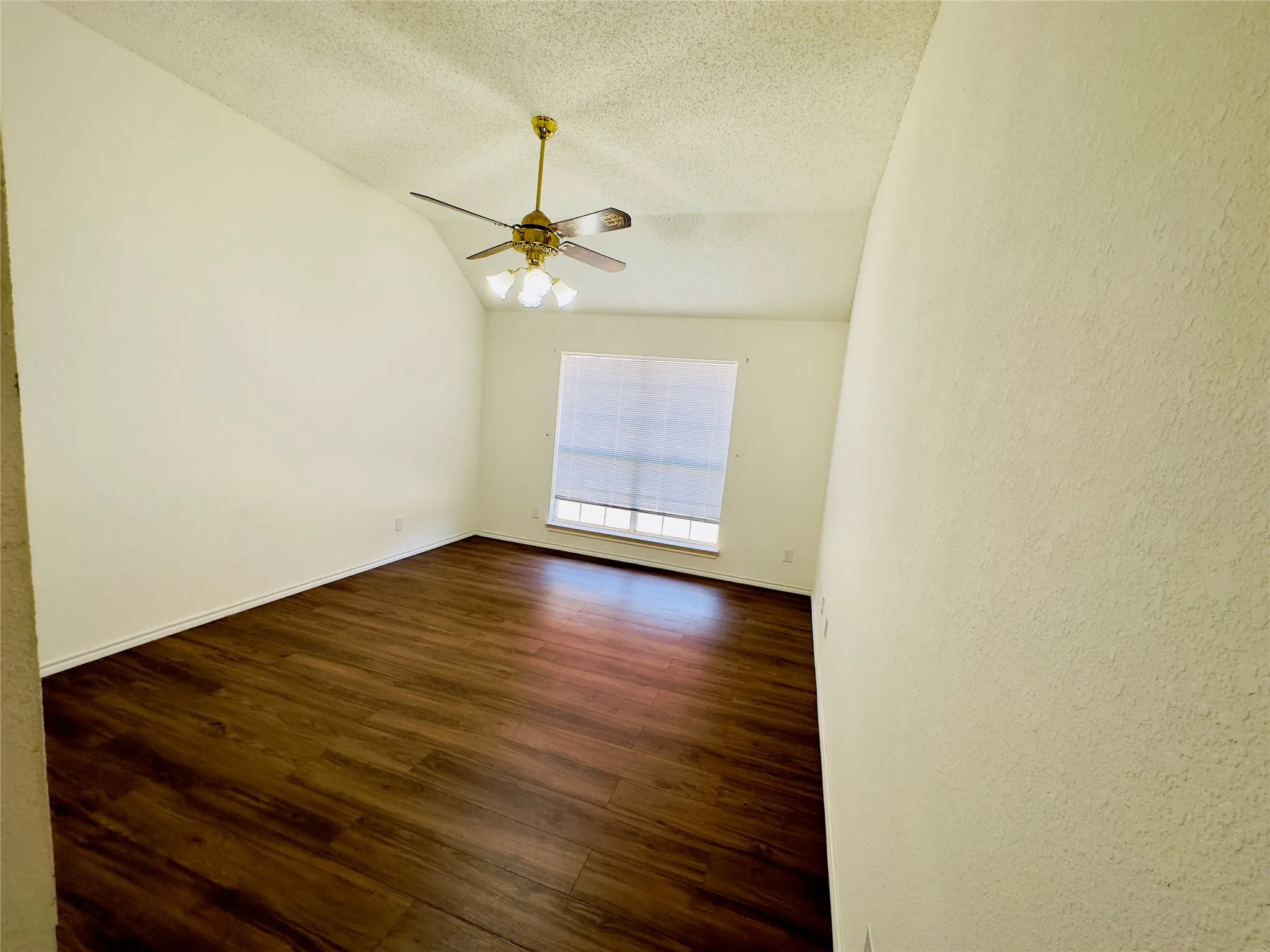 Vaulted ceiling featuring a brass-finish ceiling fan with integrated lighting, wood-finish flooring, and a window with blinds