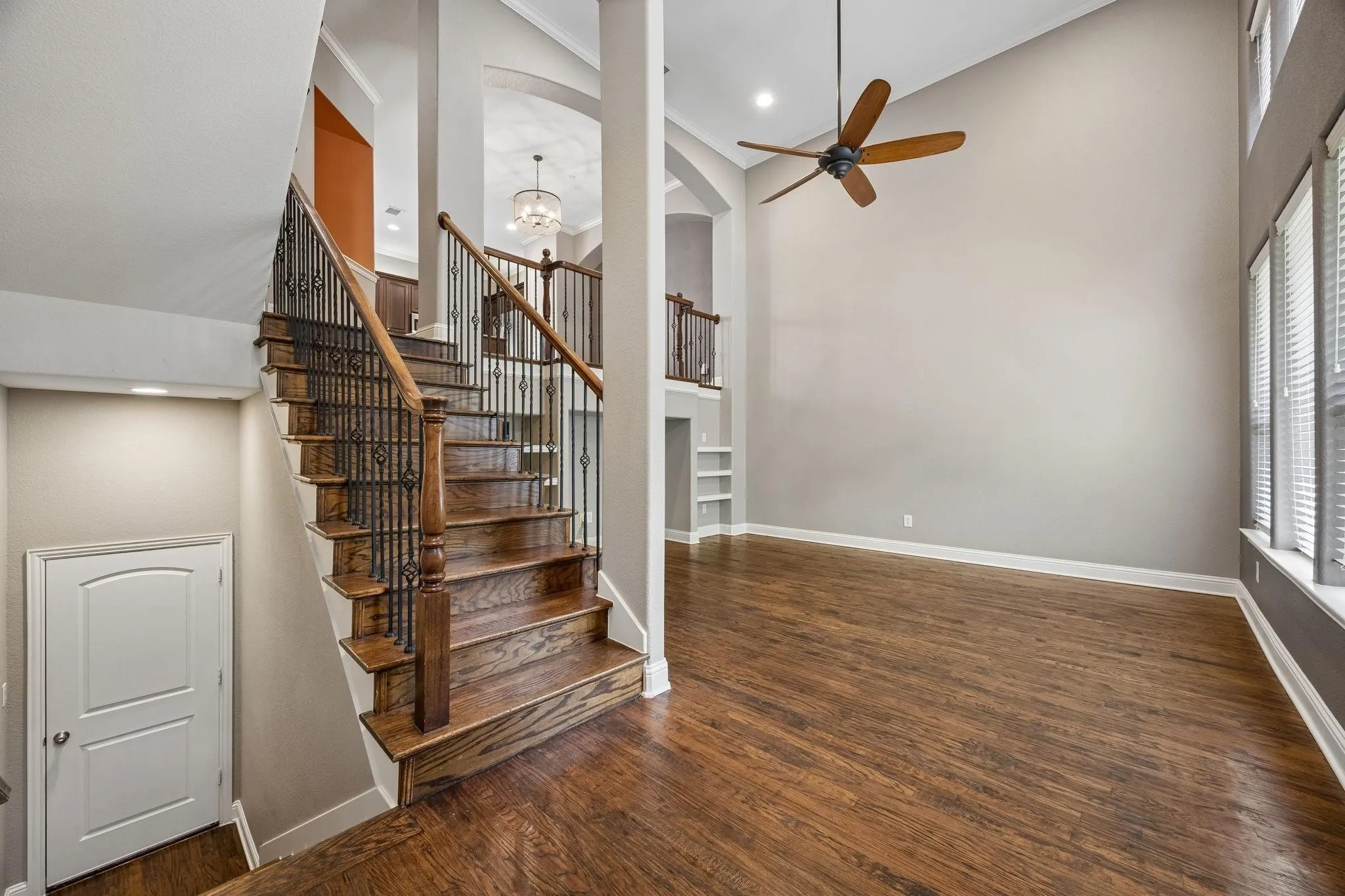 Hardwood flooring throughout, a grand staircase with wrought iron balusters, high ceilings, built-in shelving, and multiple windows with blinds